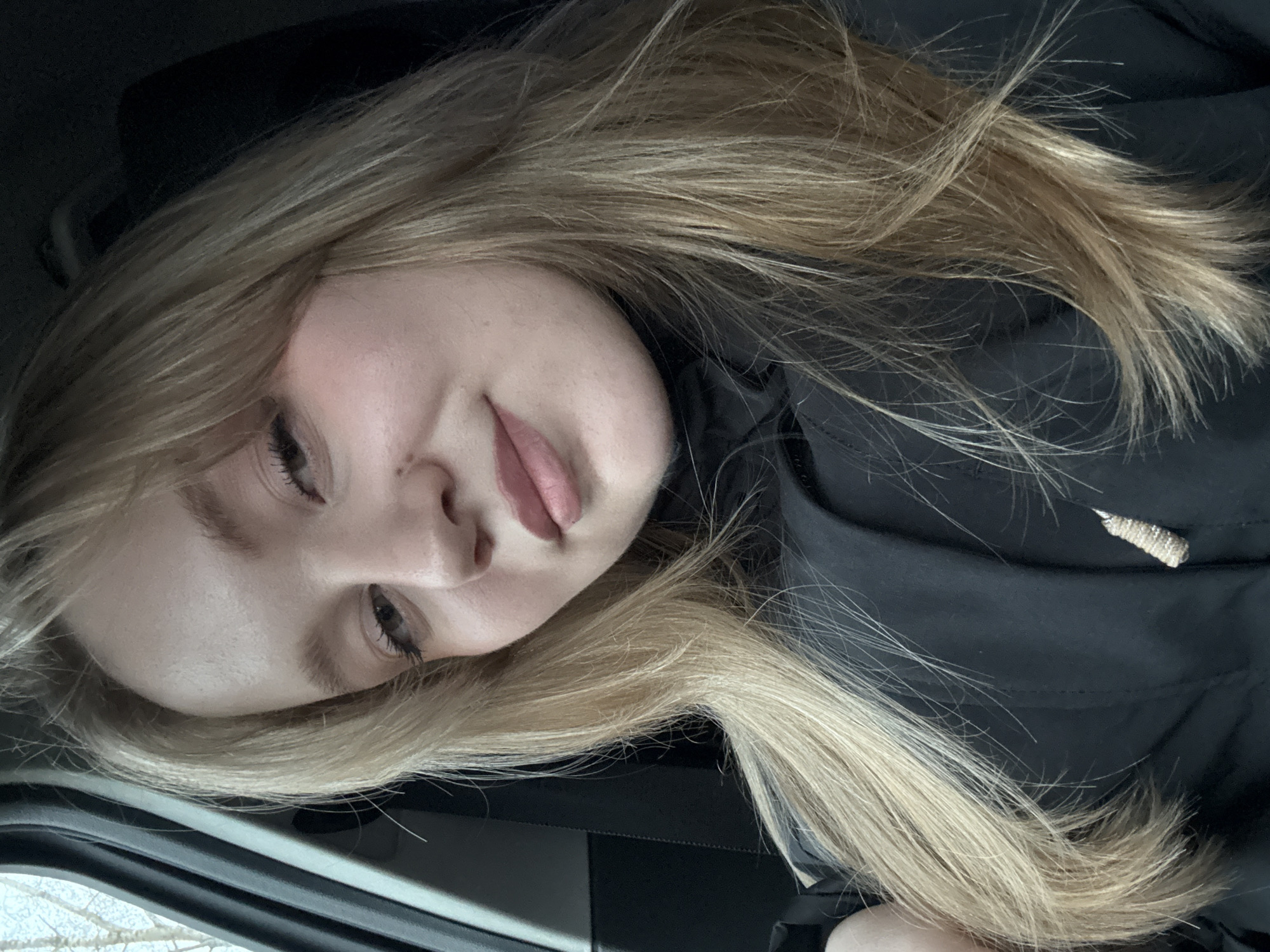 A young woman with long, light blonde hair is featured in a diagonal close-up shot, appearing relaxed and comfortable in what seems to be a car interior in Chaikovsky, Russia. Her head is tilted, and she looks subtly towards the camera with a soft, gentle expression, her lips slightly parted. Her eyes are partially visible, framed by dark eyelashes. She is wearing a black jacket or coat, with a small, textured, light-colored cord detail visible on the upper right side of her garment. The background behind her is dark, suggesting a car seat or upholstery. Through a window at the bottom left, a blurry outdoor scene with bare branches or sparse winter vegetation against a light background is visible, indicating daytime, possibly with overcast weather or snow on the ground. There is no visible text.