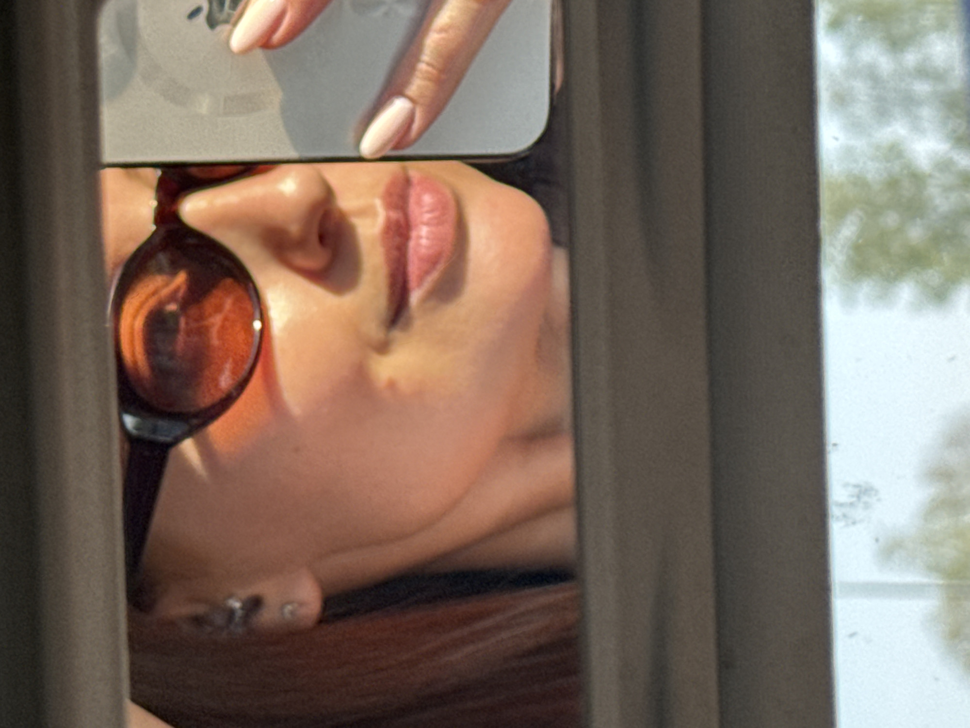 A close-up shot shows a person's face, partially obscured by a reflective surface, likely a phone held by a hand with manicured nails. The face includes a nose, lips, and an ear with piercings. Part of a round lens object is visible near the nose. The image is framed by a window frame. The background outside the window shows trees. Location information is not discernible in the provided image.