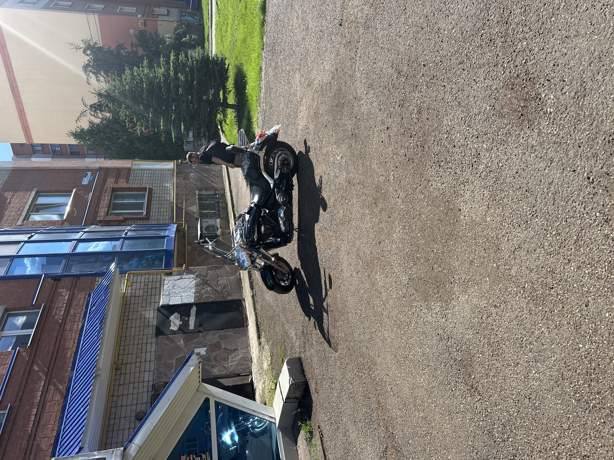 A black motorcycle is parked on a gravel lot in front of a brick building. A person wearing a dark shirt and shorts stands next to the motorcycle. The building has a blue-tinted glass facade and a blue metal roof over an entrance. A green spruce tree is visible behind the building. The scene is illuminated by bright sunlight, casting shadows on the ground. The location is likely in a residential area of Mariinsky, Russia.