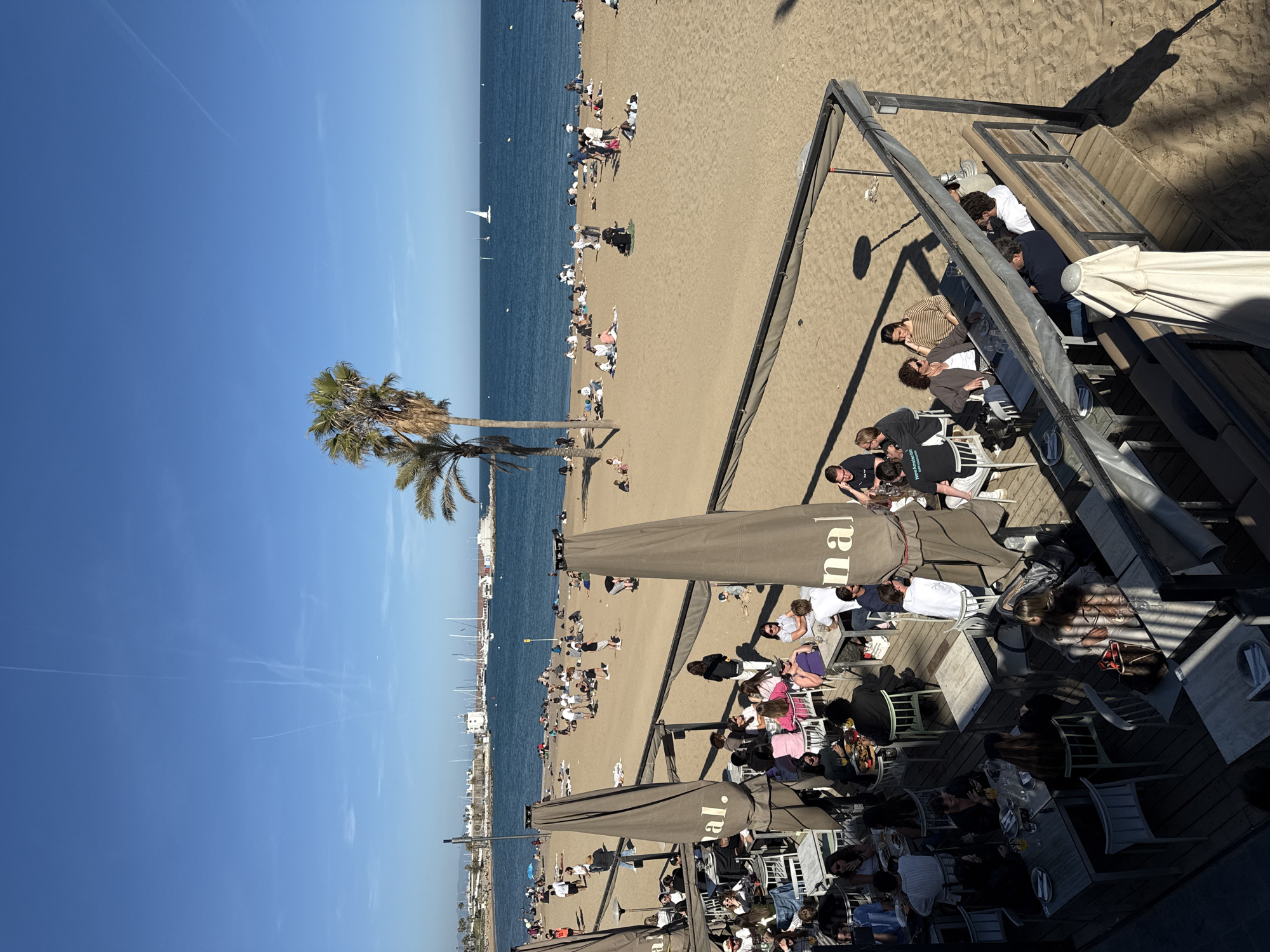 The image depicts a beach scene in Barcelona, Spain. The beach is crowded with people sunbathing, swimming, and walking. A few palm trees stand near the shoreline, and several boats are visible in the water. Adjacent to the beach is an elevated wooden terrace with multiple outdoor seating areas, accommodating many patrons enjoying food and beverages under large umbrellas. People are seated at tables, engaging in conversation and dining. The sky is clear and blue, and the sun is shining brightly.
