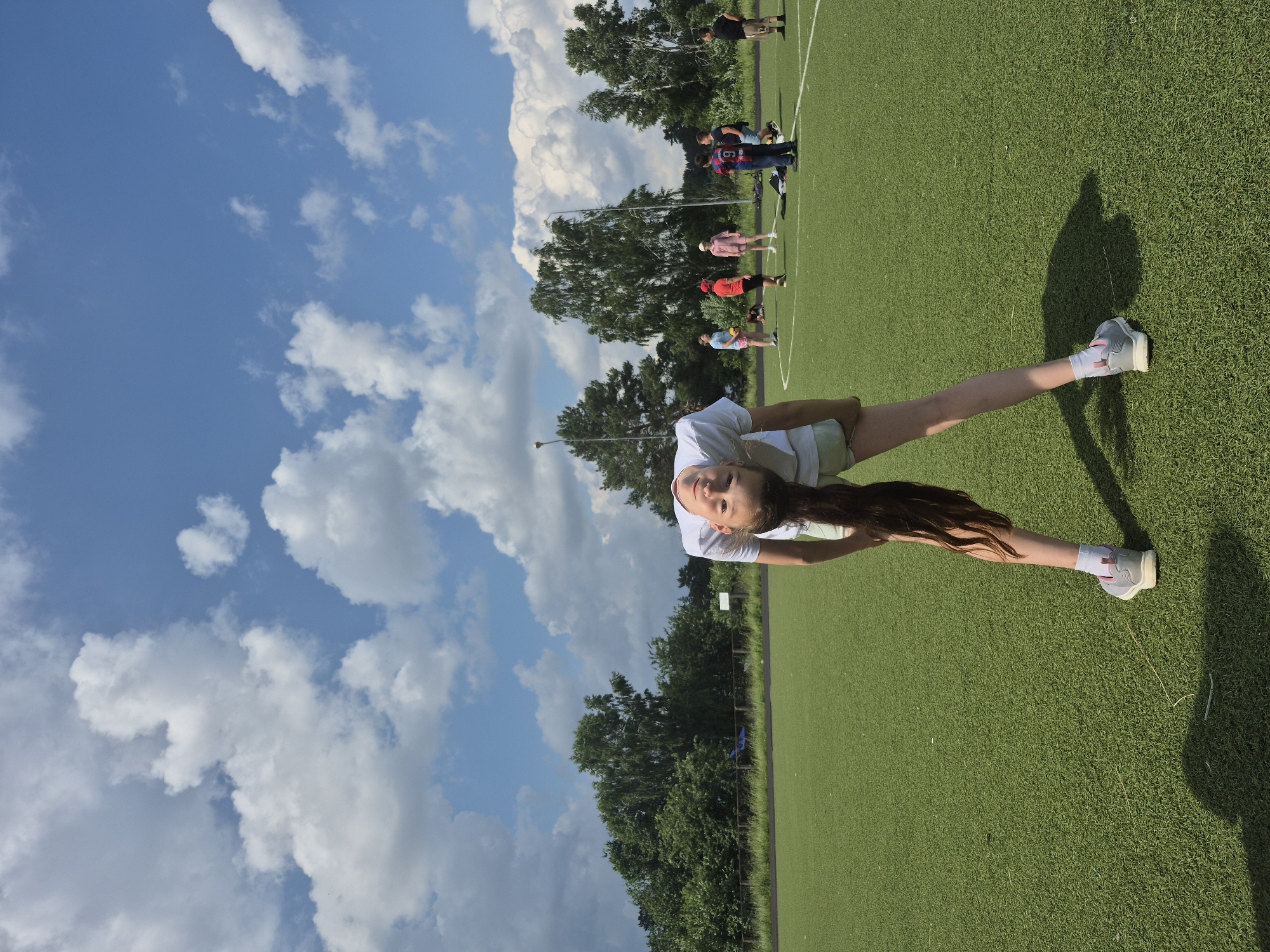 A young girl with long dark hair is performing a backbend on a green artificial turf field with white markings, possibly a sports stadium. She is wearing a white t-shirt and light-colored shorts with white socks and gray sneakers. Her body is curved backward, with her head tilted down and her hair trailing towards the ground. In the background, several other people, likely children and adults, are scattered across the field, some appearing to be engaged in activities. The environment is outdoors, under a blue sky with fluffy white clouds, and surrounded by trees. The lighting suggests it is daytime, likely a sunny day with some cloud cover. The location is identified as a sports field in Chaikovsky, Russia.