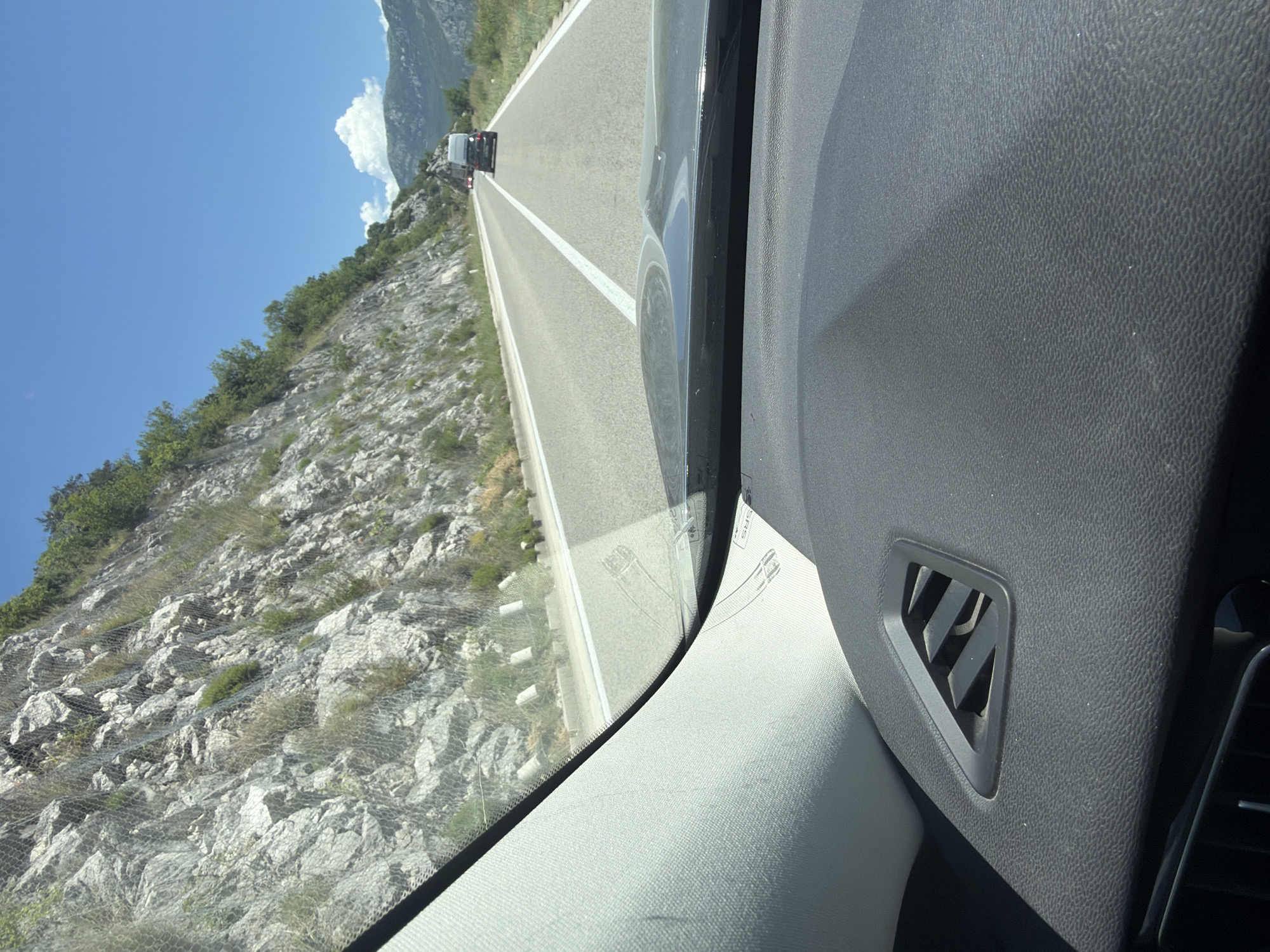 The image shows a view from inside a vehicle driving on a paved road. To the left, a steep, rocky mountainside is visible, secured with wire mesh and dotted with sparse vegetation. The road is a two-lane highway with white lines marking the lanes. In the distance, a white van is driving in the same direction as the camera vehicle, followed by a dark-colored car. The sky is clear blue with a few white clouds in the distance. The interior of the vehicle is visible in the foreground, including a dashboard with an air vent and part of the windshield. The scene appears to be located on the coastal road in Montenegro, specifically near Lipci, which is known for its scenic mountainous terrain and proximity to the Adriatic Sea.