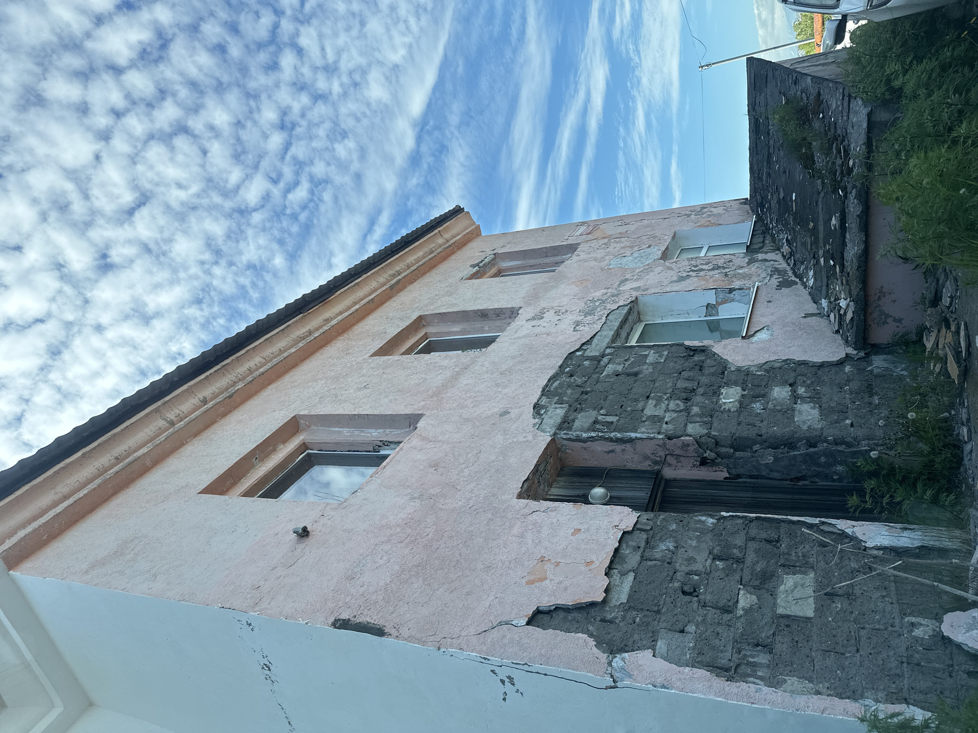 The image shows a dilapidated, pale pink building with peeling plaster and exposed brickwork. The building has several windows, some with intact glass and others broken or boarded up. There is a wooden door that appears weathered and a concrete ramp leading to an entrance. Vegetation is growing around the base of the building and on the ramp. The sky is blue with scattered white clouds. The building appears to be located in Petropavlovsk-Kamchatsky, Russia, based on its architectural style and the visible environment. The scene suggests neglect and disrepair. There are no people or animals visible in the image, and no actions or interactions are taking place. The environment is consistent with an outdoor urban setting.