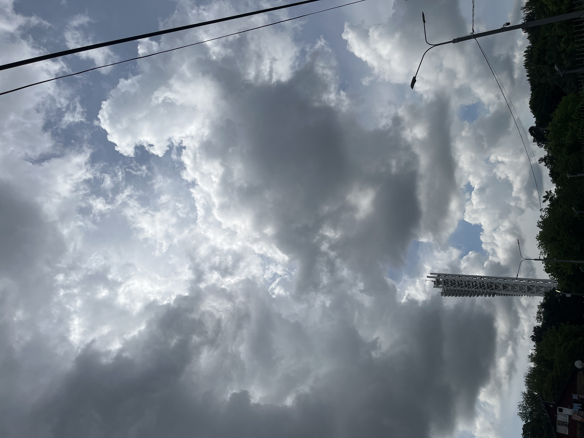 The image shows a cloudy sky above Krasnodar, Russia. Overhead power lines are visible. A tall, white lattice structure, possibly a communication tower, rises from behind trees. Streetlights are positioned on poles. A building with a visible satellite dish and windows can be seen to the right. The trees are lush and green. The clouds range from light gray to dark gray, suggesting a changing weather pattern.