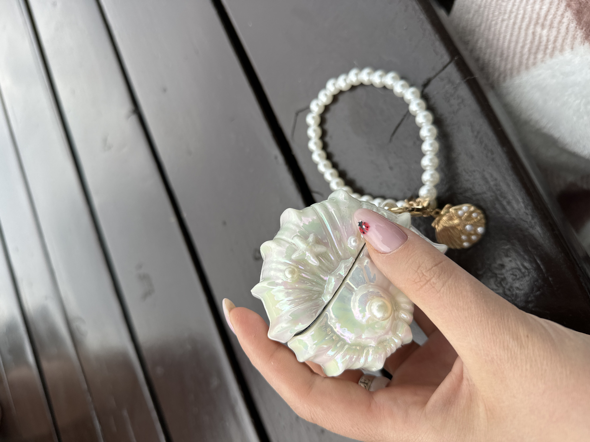 A close-up shot shows a hand holding a pearlescent shell-shaped case next to a pearl bracelet on a dark wooden surface. The background includes a blurred white and beige plaid pattern. The scene does not provide any information about the location in the city of Šušanj, Montenegro.