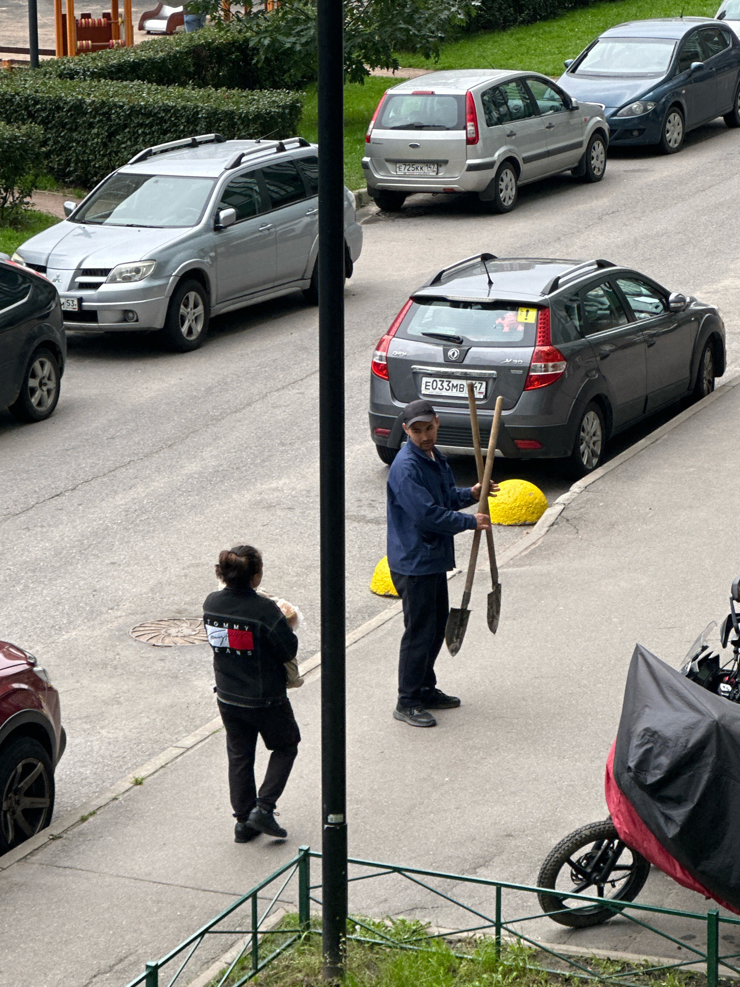 In Murino, Russia, on a paved street, a man wearing a dark blue jacket and black pants stands holding two shovels. He has a dark cap on his head and appears to be looking towards the right of the frame. He is standing next to a grey hatchback car. To his left, a woman wearing a black jacket with a Tommy Hilfiger logo on the back is seen from behind, holding a light-colored object. She is wearing black pants and dark shoes. Several cars are parked along the street, including a silver SUV, a dark blue sedan, and a silver hatchback. In the background, there is a neatly trimmed hedge and a playground structure. A partially covered motorcycle is visible in the lower right corner of the image.