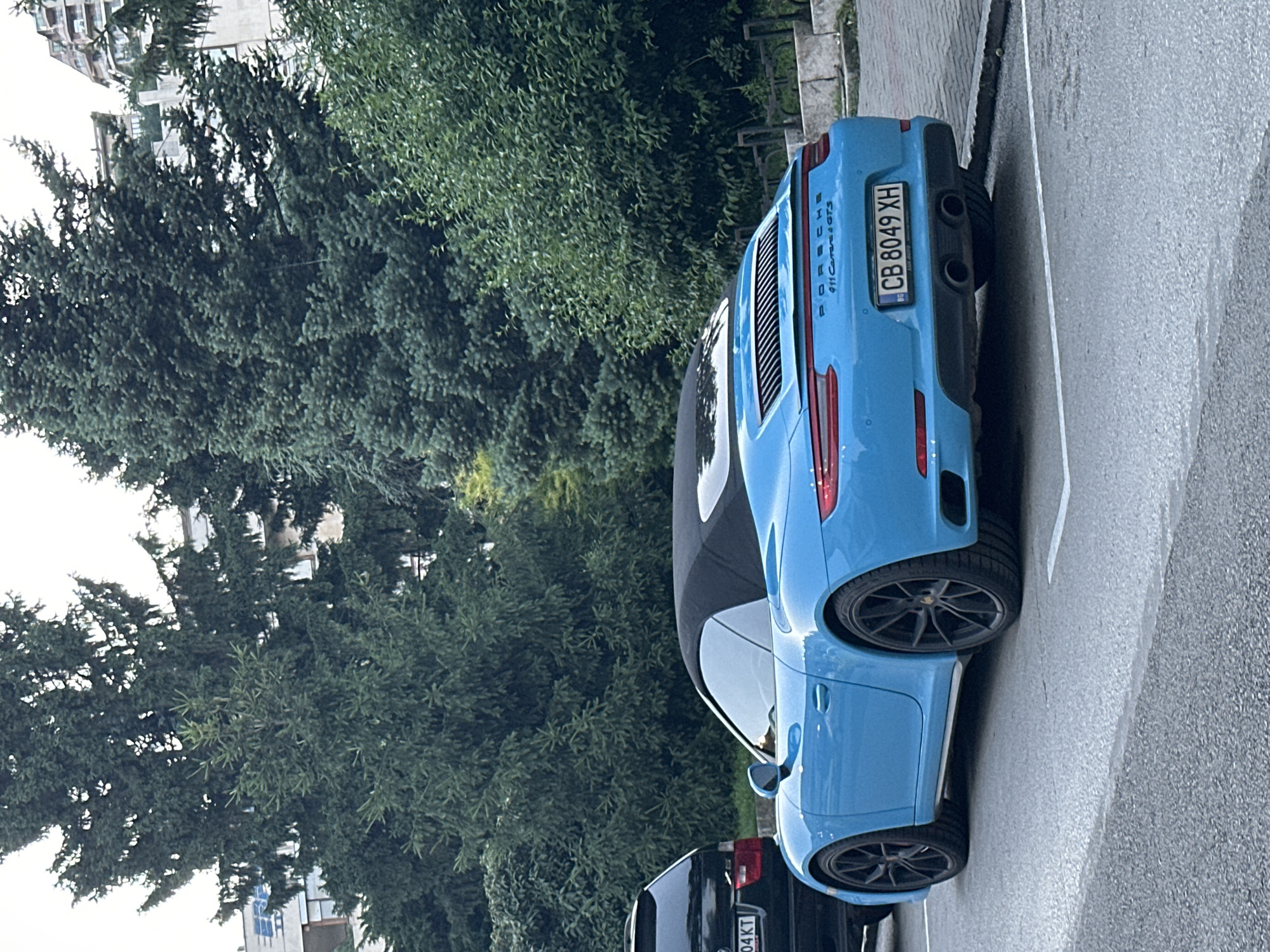 The image shows the rear and side view of a bright blue Porsche 911 Carrera 4 GTS convertible parked on a street in Veliko Tarnovo, Bulgaria. The car has a black soft top. Its license plate reads "CB 8049 XH". To the left of the Porsche, a dark-colored SUV is partially visible, with its rear bumper and license plate showing "04 KT". The cars are parked on an asphalt surface marked with white parking lines. In the background, lush green trees and a building with balconies are visible. The overall setting appears to be an urban environment with a focus on the vehicles.