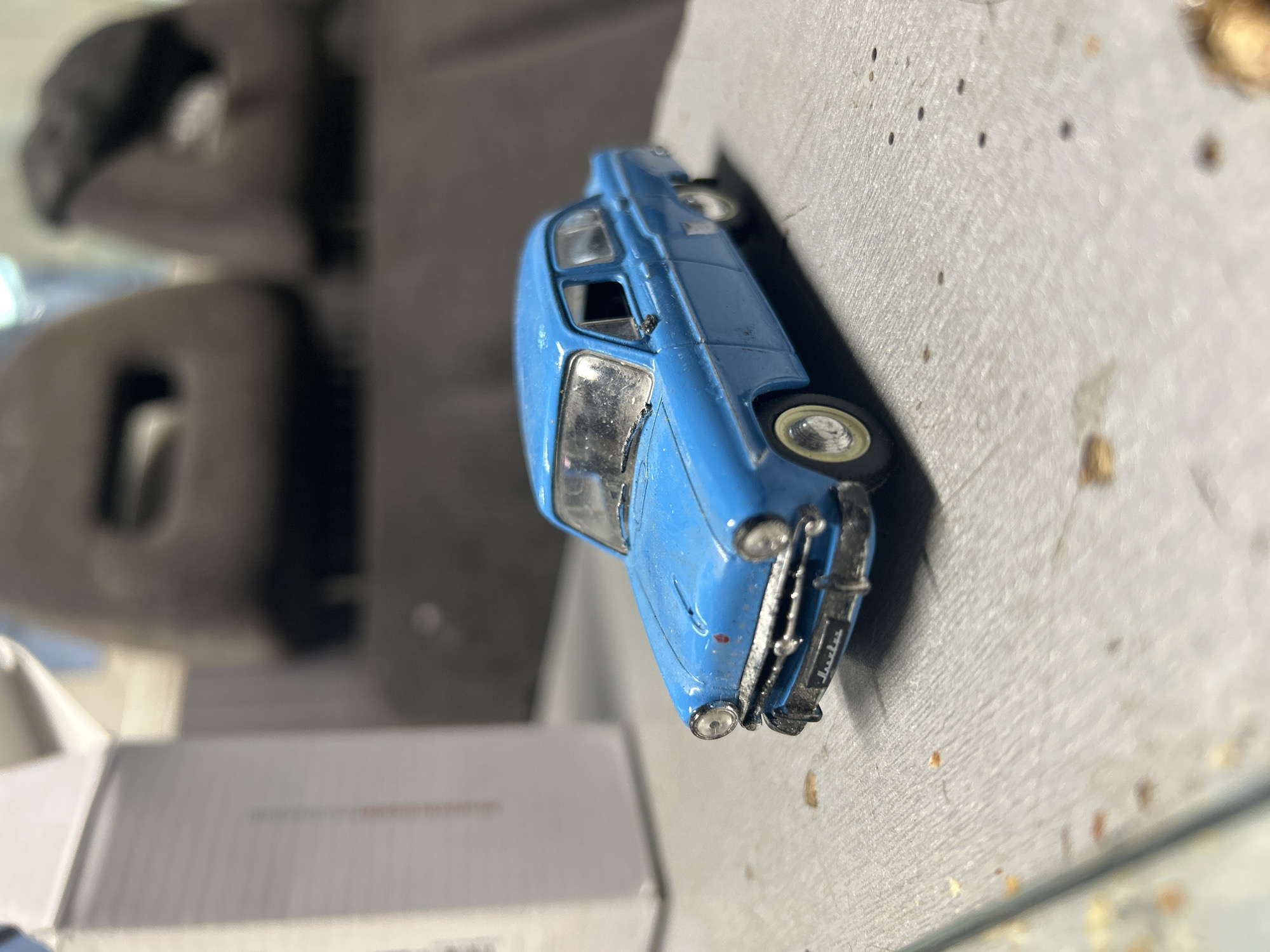 A close-up shot presents a blue model car, possibly a "Trabant", resting on a gray surface. The car is positioned at an oblique angle, with the front of the car facing the upper left of the frame. In the background, out of focus, are gray objects and a white box. The location is unknown.