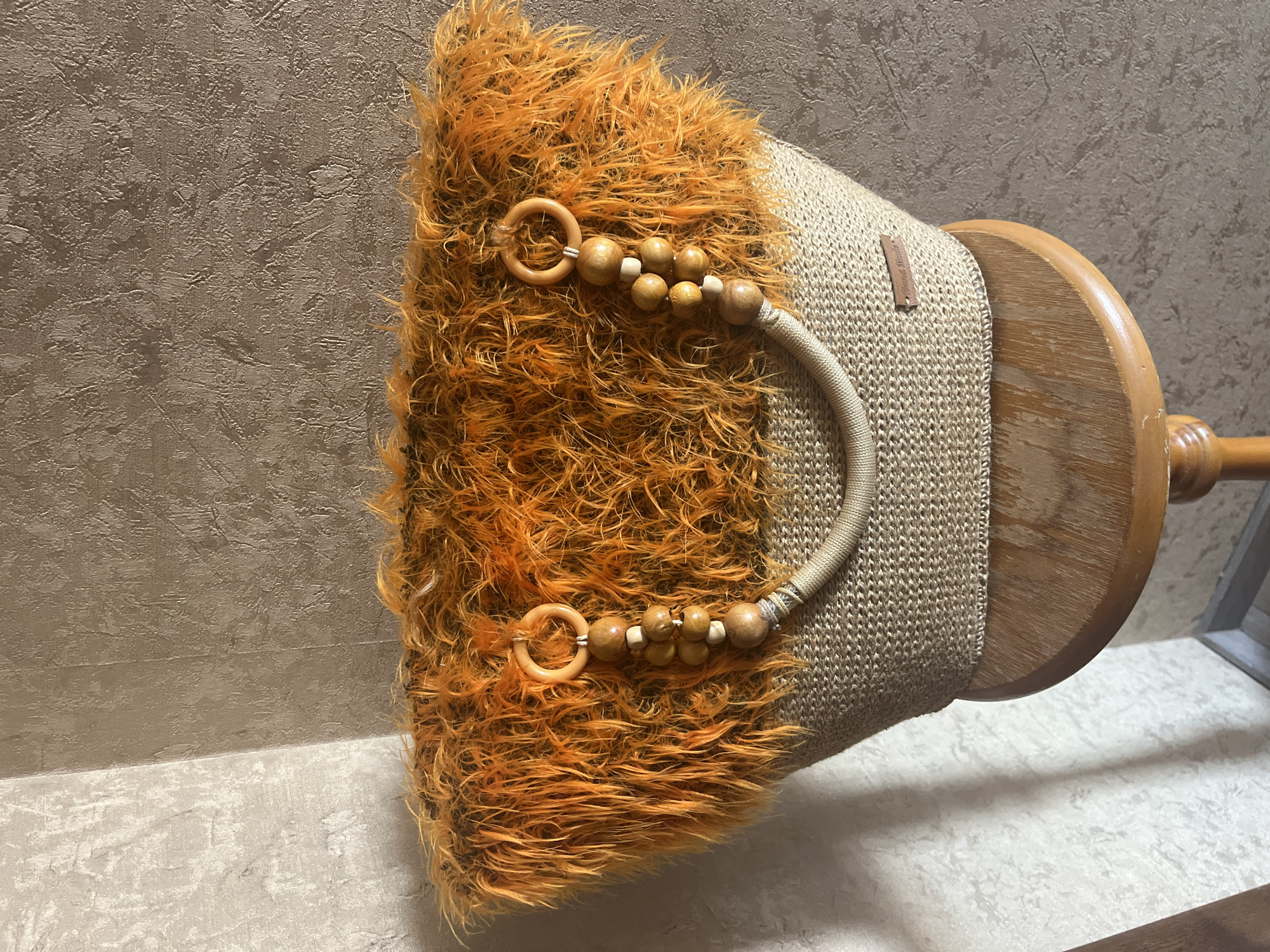 The image shows a close-up of a unique, handmade tote bag. The bag has a woven raffia or straw body in a neutral beige color. The top portion of the bag is adorned with fluffy, bright orange faux fur. Attached to the bag are two rounded wooden handles, each decorated with a strand of wooden beads of varying sizes. A braided rope detail connects the two handles across the front of the bag. On the lower right side of the bag's body, there is a small, rectangular brown tag, possibly made of leather, with embossed text that appears to say "Hand Made". The bag is resting on a round wooden stool, which has a worn, polished finish and is supported by a central pedestal base. The background consists of textured walls, one in a light gray with a subtle pattern and the other in a darker, mottled brown. The lighting suggests an indoor setting with natural light casting shadows. There is no indication of the city of Vladimirovka, Russia, in the image or its context.