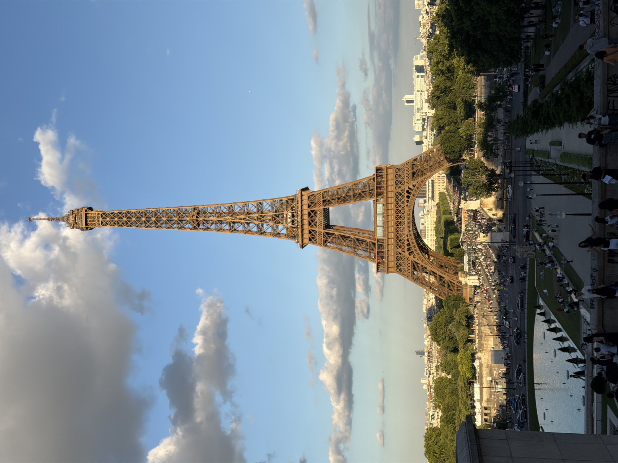 This media file captures a view of the Eiffel Tower in Paris, France, during what appears to be late afternoon or early evening. The sun is casting a warm glow, suggesting favorable weather with a mix of blue sky and scattered clouds.
The primary subject is the iconic Eiffel Tower, with its intricate latticework structure dominating the left side of the frame. The tower is viewed from a lower vantage point, looking up towards its peak.
Below and to the right of the tower, a bustling scene unfolds. Numerous people are visible, scattered across pathways, grassy areas, and near a body of water which could be a reflecting pool or a part of the Seine River. Some individuals are standing, others are seated, and a few are walking. Cars and other vehicles are present on a road or pathway adjacent to the park-like setting. The presence of many people suggests a popular tourist spot or a public gathering.
Beyond the immediate area around the tower, a cityscape is visible, characterized by numerous buildings, some of which appear to be apartments or offices. Lush green trees line the pathways and parks, adding to the urban landscape. The overall mood seems relaxed and lively, indicative of a pleasant day in a well-visited tourist destination. No visible text is present in the image.
