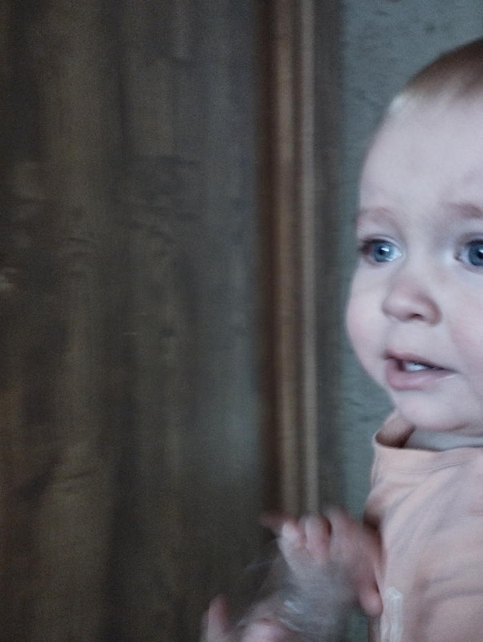 A toddler with blue eyes and light hair is the focus. The child looks toward the left side with a slightly open mouth. The background consists of a blurry brown wall, potentially made of wood, with vertical lines or patterns visible. The toddler is wearing a light-colored shirt. One of their hands is visible with fingers slightly curled.