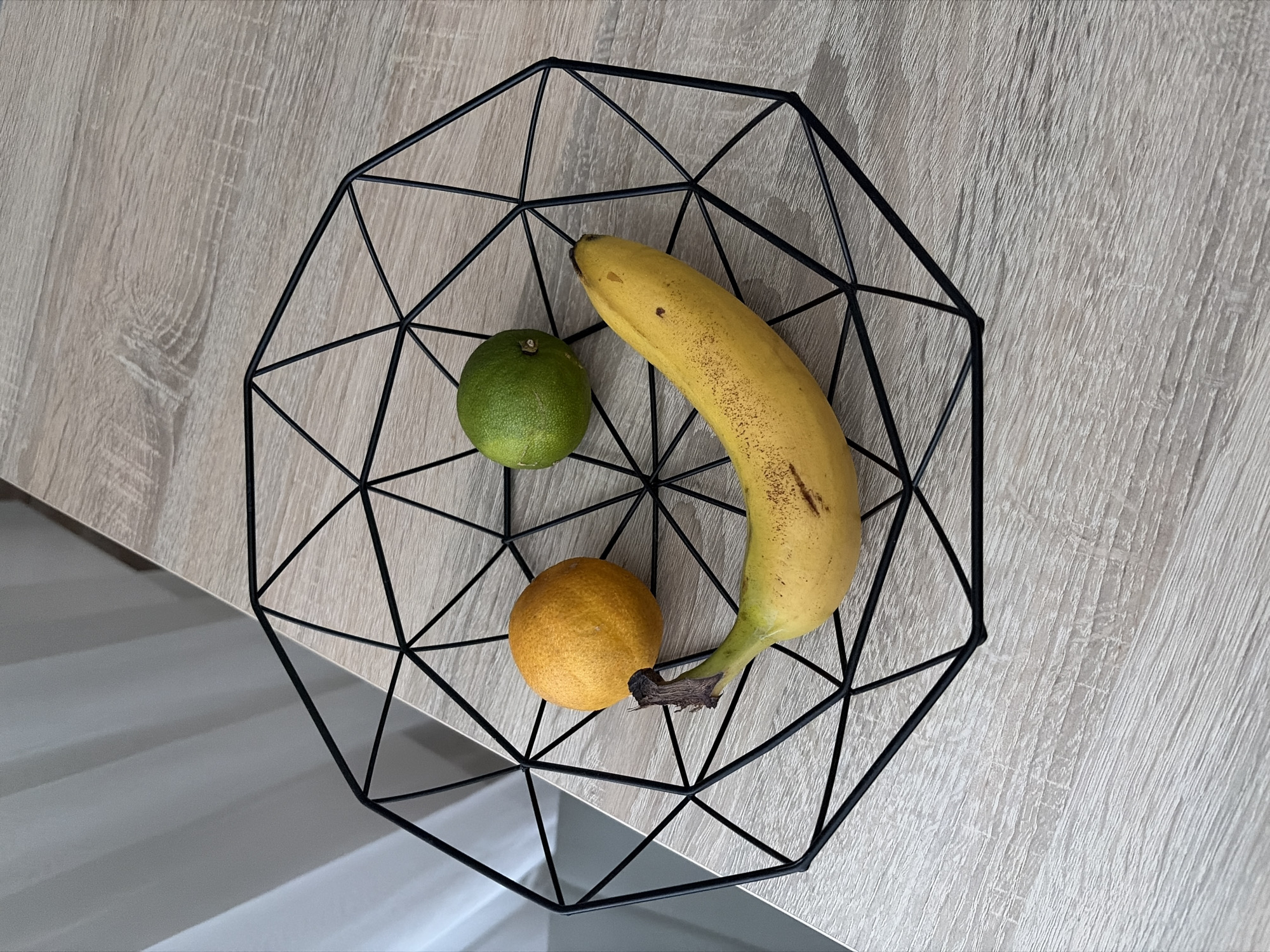 A fruit basket made of black wire, arranged in a geometric pattern, contains a banana, a lime, and an orange. The basket is placed on a light-colored wooden surface, possibly a table. In the background, a white curtain is visible. The lighting suggests an indoor setting. The overall composition creates a visually appealing arrangement of the fruits within the modern basket. There are no people or animals in the image, and no specific location in Nitra, Slovakia can be identified from the visual information provided.