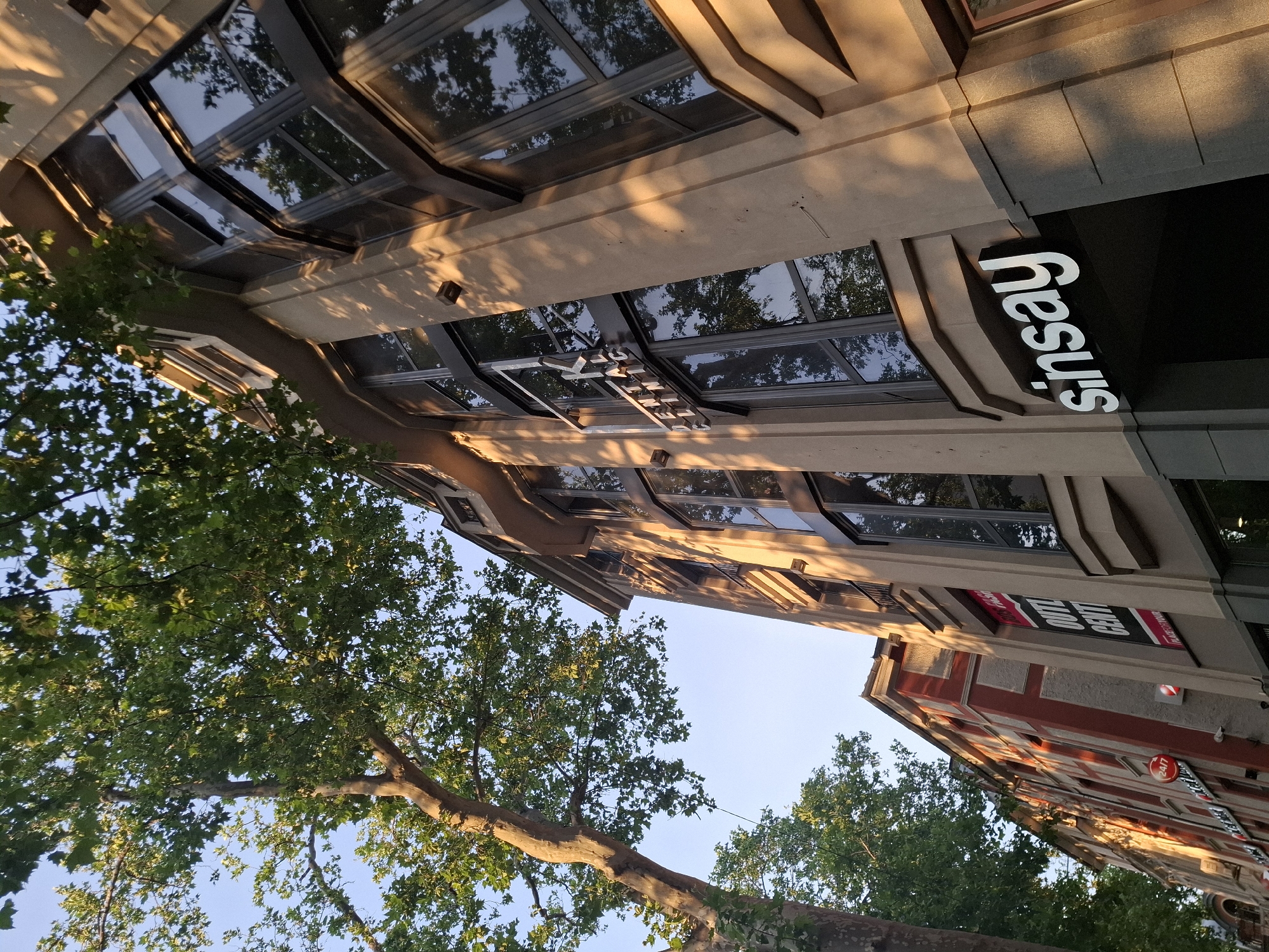 A low-angle outdoor shot captures a building with large windows, reflecting trees and the sky. The building's facade is a light tan color with darker grey trim around the windows. One prominent sign reads "sinsay" in white, raised lettering. To the left, another building with red and white accents is partially visible, also featuring large windows. A large tree with green leaves is in the foreground, casting shadows on the building. The sky is clear and blue.