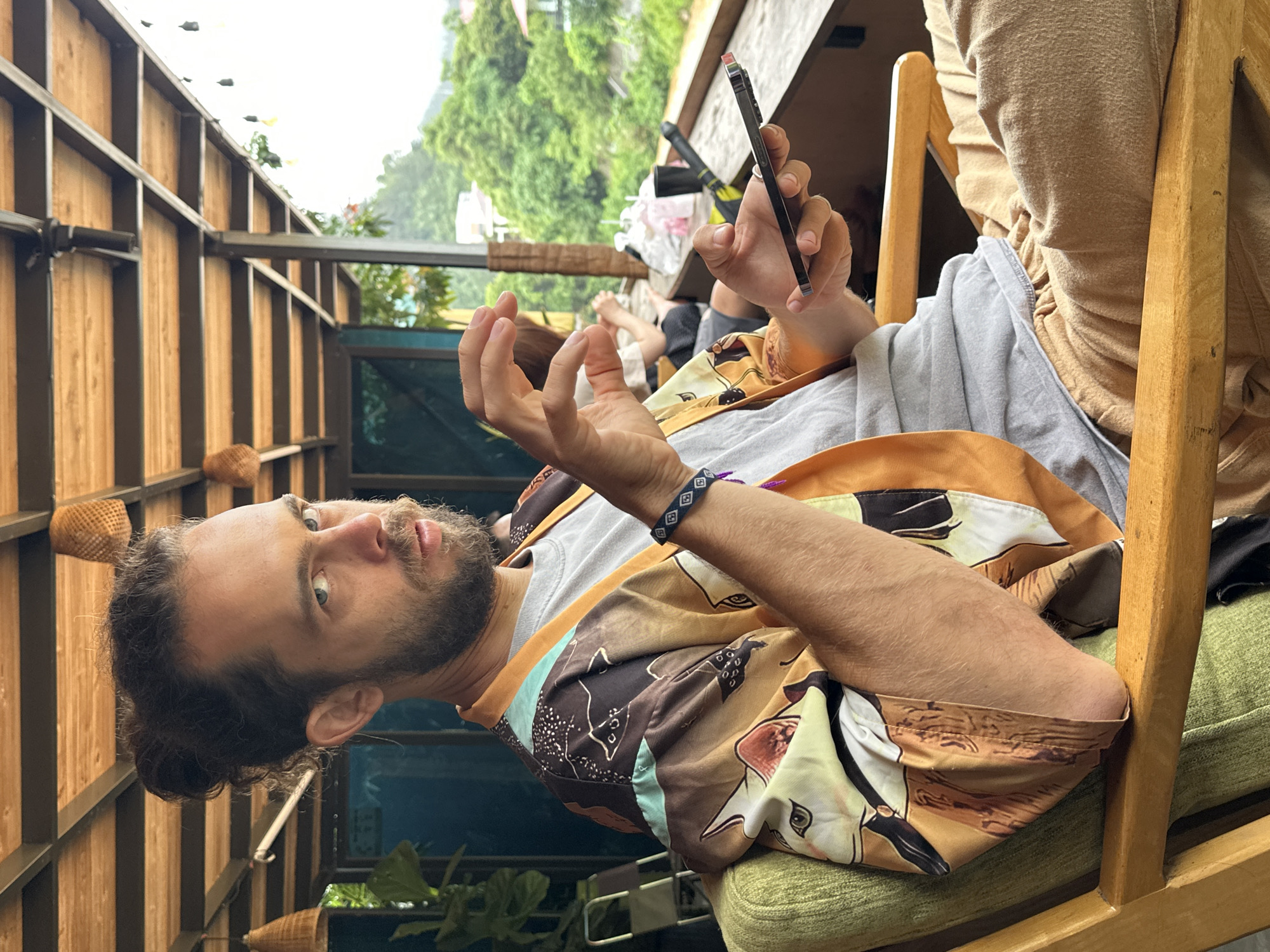 A man with a beard and dark hair pulled back into a bun is seated on a wooden chair with green cushions, gazing directly at the viewer. He wears a grey t-shirt underneath a vibrant, patterned button-up shirt featuring animal motifs in shades of orange, brown, and light blue. A blue and white woven bracelet adorns his left wrist. His left hand is raised in a gestural pose, while his right hand holds a dark-colored smartphone upright.
The setting appears to be an outdoor or semi-outdoor space, possibly a cafe or guesthouse balcony. To the man's left and behind him is a wooden slatted wall, interspersed with dark metal framing elements. Small, woven straw baskets hang decoratively from this wall. In the background, lush green trees and foliage dominate the landscape under a bright, clear sky, indicating a natural, perhaps mountainous or hilly environment. The legs of another person, possibly resting on a cushion, are partially visible in the mid-ground.
The scene unfolds during the daytime under clear weather conditions. The rustic wooden architecture and the verdant, expansive backdrop are strong indicators of a scenic, natural location like Mù Cang Chải, Vietnam. The man’s expression is calm and engaged. No visible text is present in the image.