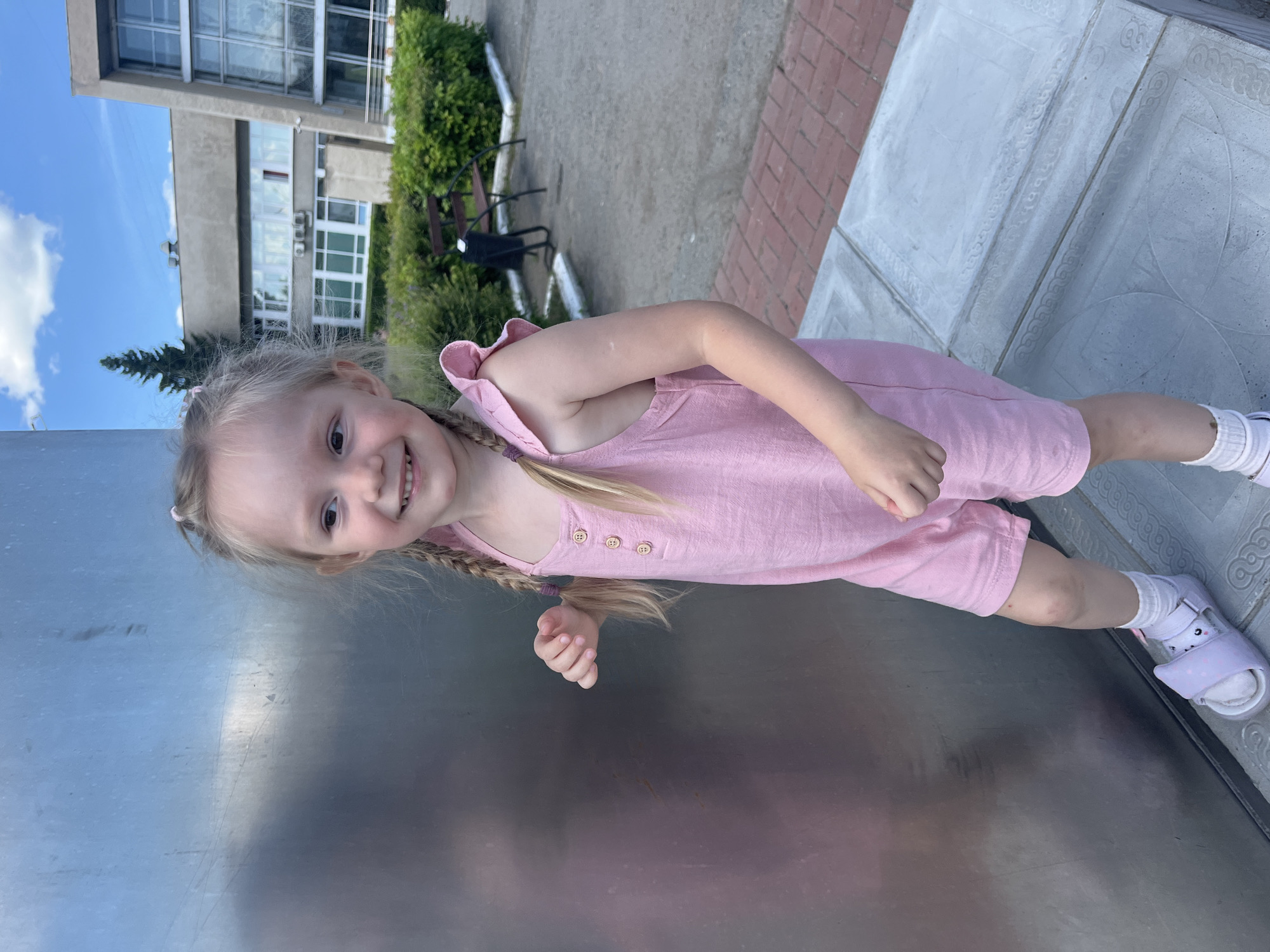 A young blonde girl with pigtails, wearing a pink romper and white socks with sandals, stands in front of a large, metallic structure in Shadrinsk, Russia. The girl is smiling and looking to her left. Behind her, a modern building with large windows and a tall evergreen tree are visible. The ground is paved with concrete slabs and bricks, and there are bushes and a bench in the background. The sky is blue with scattered clouds.