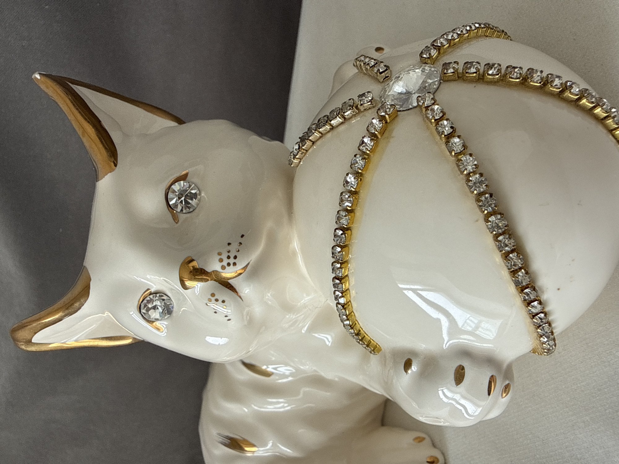 A porcelain cat figurine is shown in a close-up shot. The cat is white with gold trim on its ears, nose, and whiskers. Two small gemstones adorn the eyes on the cat's face. On the cat's back, a ball is placed, decorated with a diamond and rhinestone-studded bands. The background is a neutral gray and white color. The location of Votkinsk, Russia is not evident in this image.