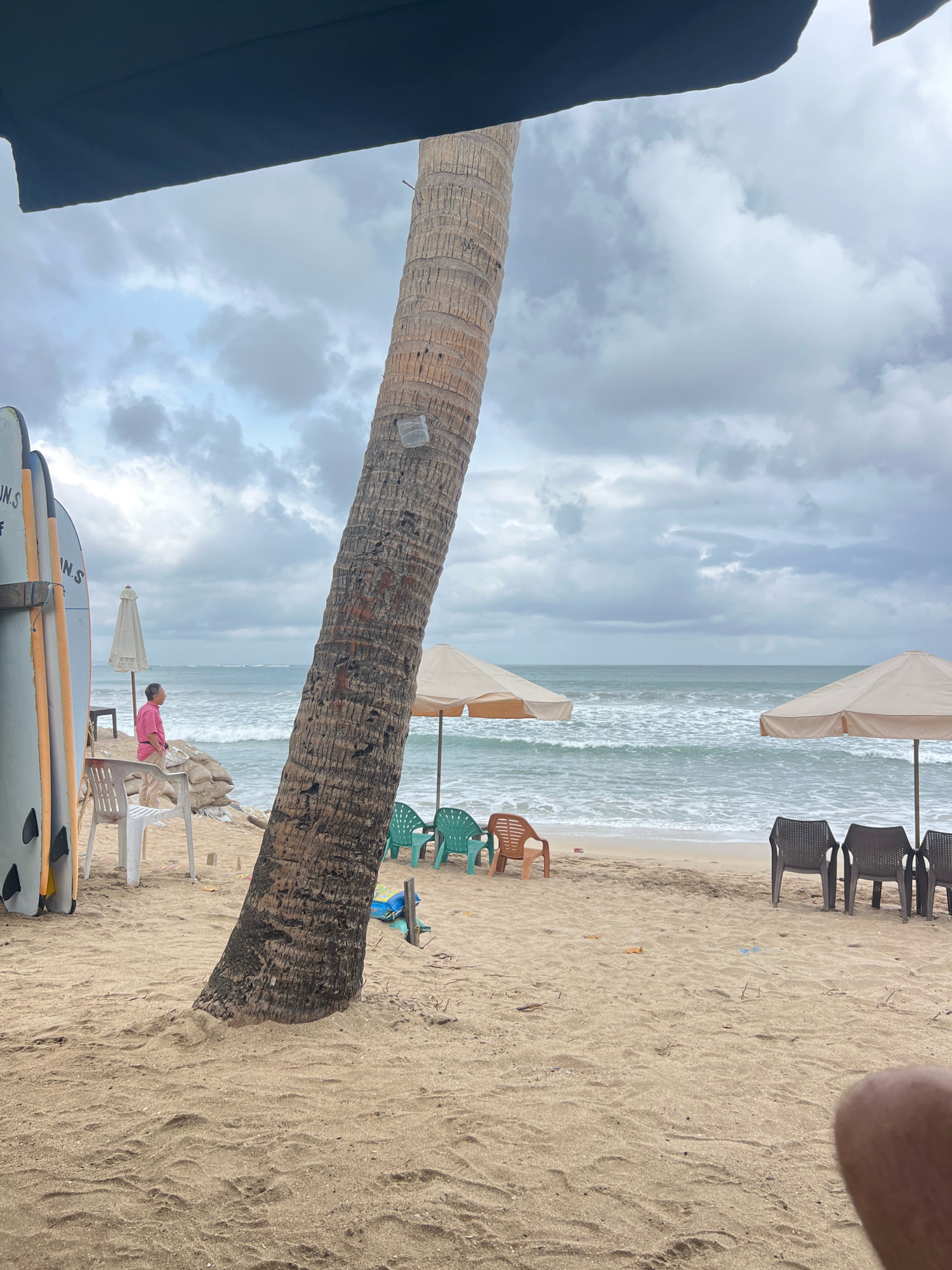 The image depicts a beach scene in Kuta, Indonesia, under a cloudy sky. In the foreground, a large, weathered palm tree trunk leans diagonally across the frame, partially obscuring a dark canopy overhead. The sandy beach is sparsely populated with several chairs, some plastic and some wicker, as well as two beige beach umbrellas. A few surfboards stand upright on the left side of the image, next to a person in a pink shirt looking out at the ocean. The ocean itself is a pale blue-green with moderate waves, and the horizon is hazy. Scattered debris, including plastic and food wrappers, is visible on the sand. The overall atmosphere is subdued, likely due to the overcast weather.
