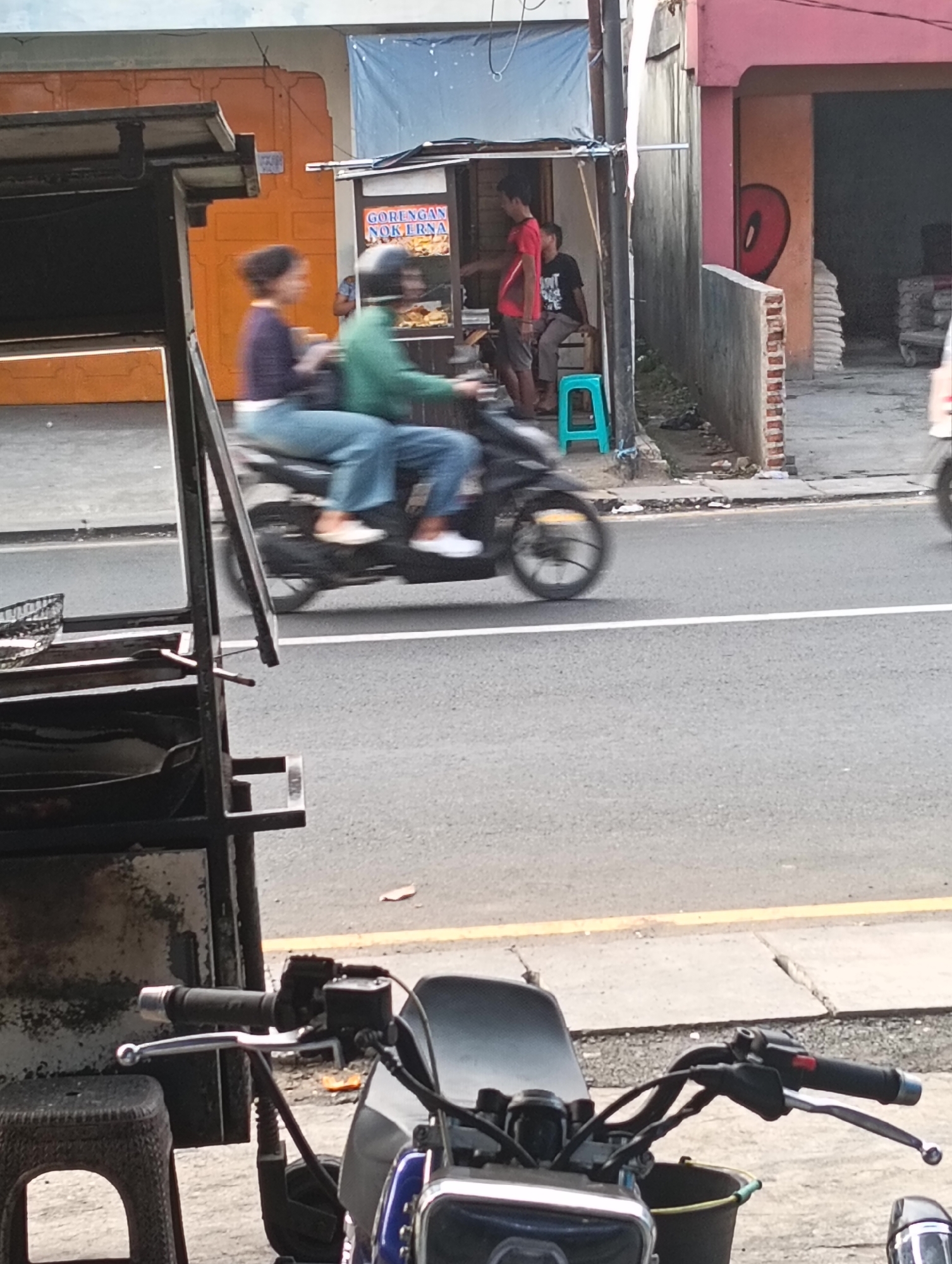 A low-angle, slightly blurry, medium shot captures a street scene in Desa Cipakat, Indonesia. In the foreground, the handlebars and front of a blue and silver motorcycle are visible, with a black plastic stool to the left. A food stall with a black metal frame and an orange corrugated metal roof is also partially in view on the left. The middle ground features a motorcycle in motion, ridden by a person wearing a green jacket and helmet, with a second person behind them wearing a purple top and blue jeans. They are traveling on an asphalt road with white and yellow painted lines. In the background, a small building with a pink facade and a grey concrete wall is visible, with a red graffiti heart-shaped design on the wall. A blue plastic stool is outside the building, and two men are standing near a food stall with a sign that reads "GORENGAN NOK ERNA". Piles of white sacks are stacked in an open doorway to the right. The sky is overcast and light.