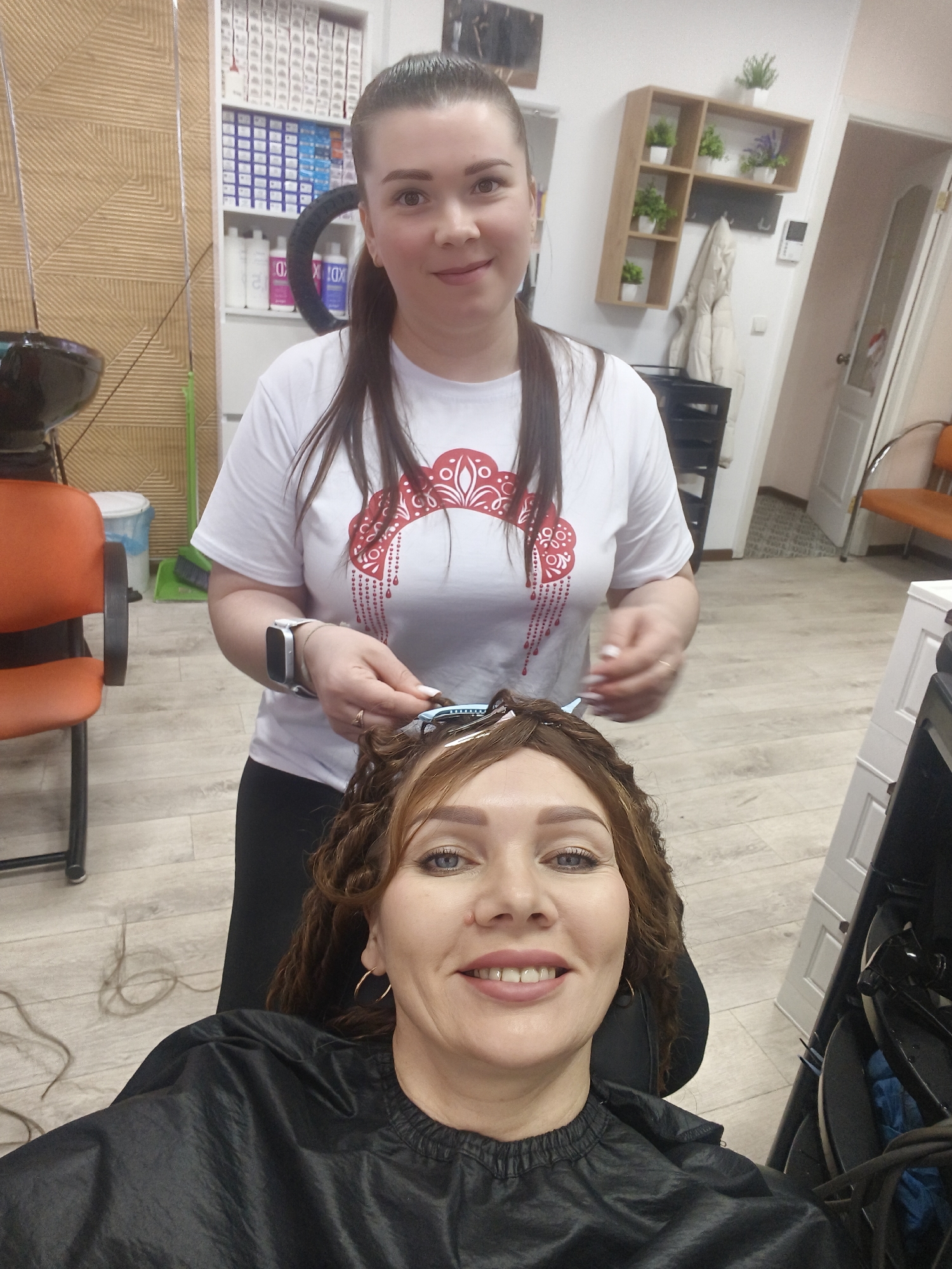 The image depicts two women inside a hair salon. One woman is seated and facing the camera, wearing a black cape and hoop earrings. She is smiling and has hair clips in her hair. The other woman, standing behind her, appears to be a hair stylist working on the seated woman's hair. The stylist is wearing a white t-shirt with a red design and a smart watch. The salon background includes a washing station with an orange chair, product shelves, a decorative wall panel, and a wooden shelf with plants.