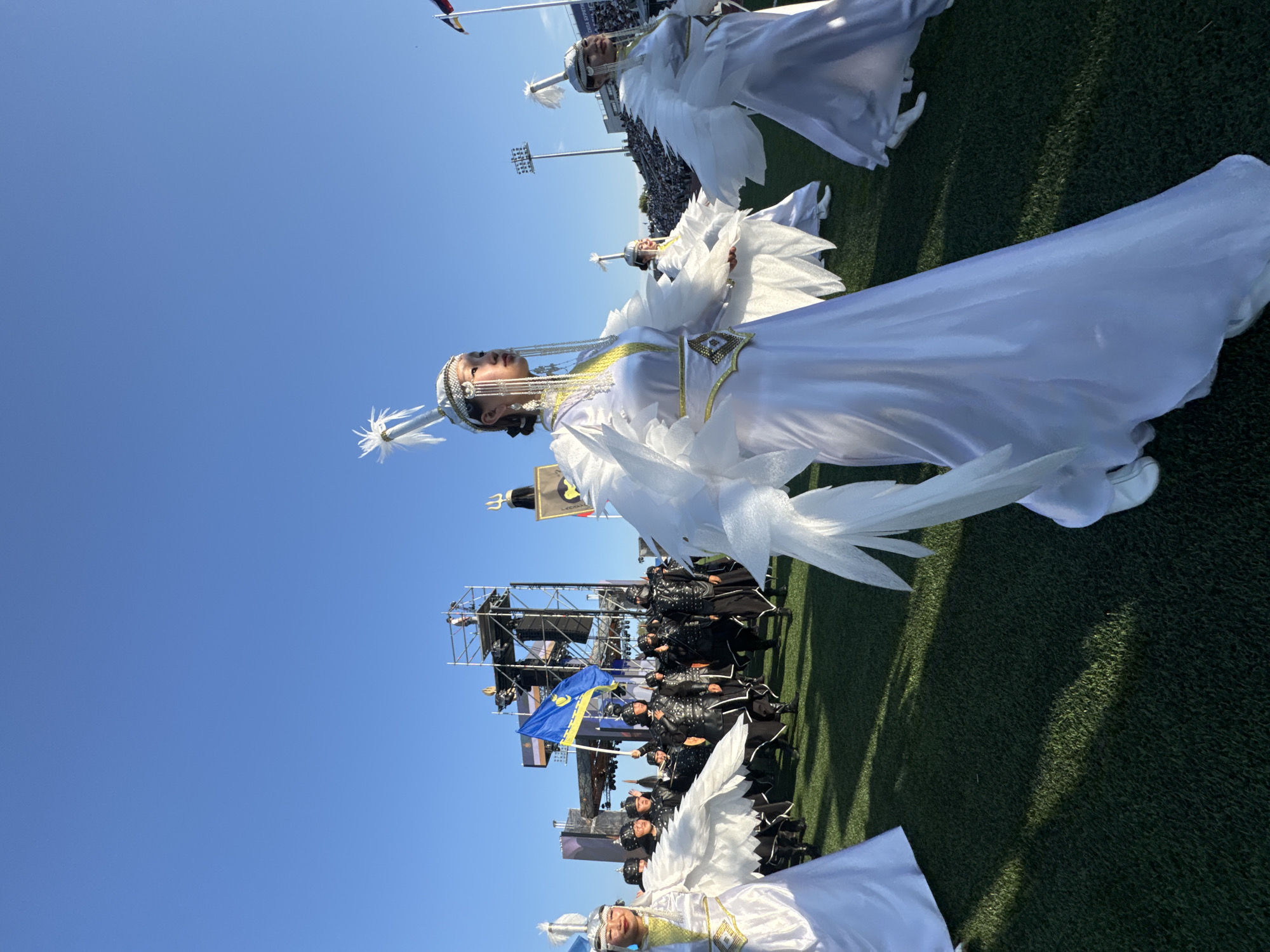 In Ulan-Ude, Russia, a group of people are participating in an outdoor event on a grassy field, likely a stadium, under a clear blue sky. In the foreground, three individuals dressed in white, flowing costumes with elaborate headdresses are dancing or posing. These costumes feature white feathery wing-like embellishments on the arms and are adorned with decorative silver and gold elements. Behind them, a larger group of people are dressed in dark, leather-like armor and helmets, appearing to be in formation or marching. One person in this group holds a flag with yellow and blue colors, featuring a blue symbol resembling a wheel with spokes. In the background, there are tall scaffolding structures, stage equipment, and stadium seating visible, suggesting a performance or ceremony is taking place. The lighting indicates it is daytime, with shadows cast across the field.