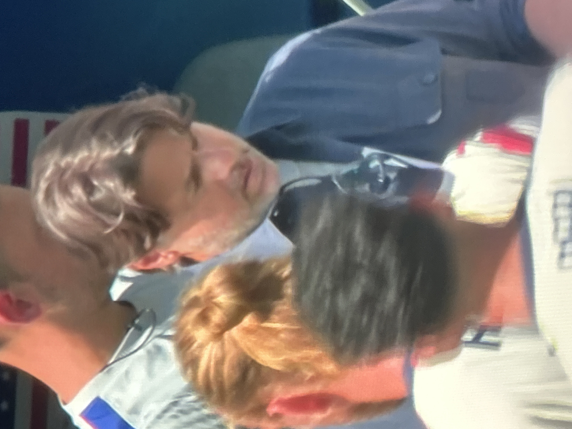 In an outdoor, possibly urban setting under a bright sky, a group of people are seen. The central figure is a man with light brown, medium-length hair, wearing a dark blue collared shirt over a gray shirt. He is looking down and to his right, with his mouth slightly open. To his left, a person with blonde hair tied back is visible from the neck up, also looking down. In the foreground, a person with dark, short hair is mostly obscured, seen from the back. In the background, another person, wearing a white shirt with blue accents, is visible from the shoulder up, with their head turned away. The overall impression is of a casual gathering.