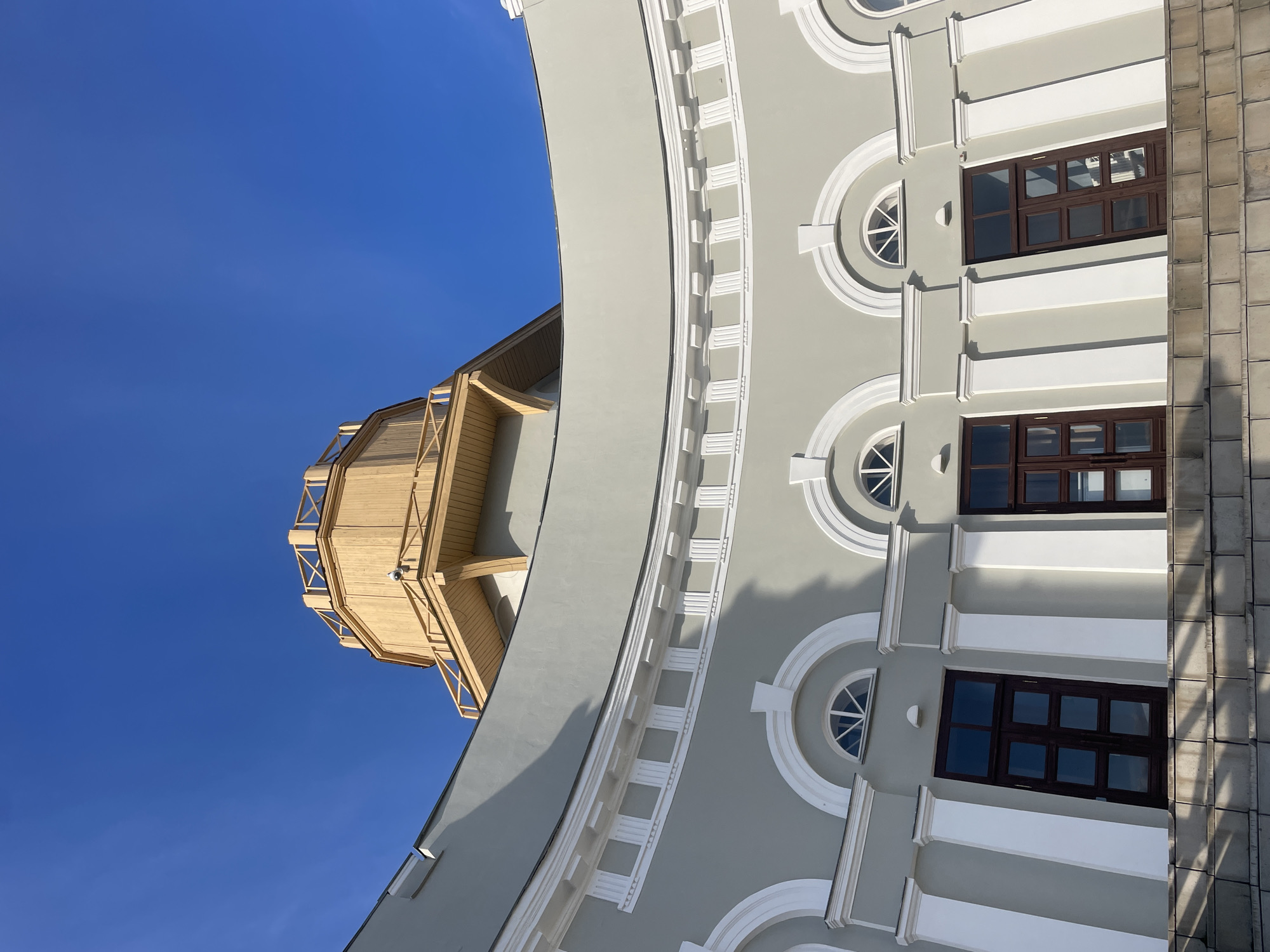 The image presents a striking architectural view against a vibrant, clear blue sky. A prominent building with a light grey, curved facade dominates the scene, extending from the lower part of the frame upwards and towards the right. This facade is intricately adorned with white decorative trim, including horizontal bands and vertical pilaster-like elements that create a sense of depth and classical style. Integrated into this design are various windows: some are semicircular with radiating white spokes, while others are rectangular with dark brown frames, reflecting the bright sky.
Emerging from behind the main grey building on the upper left side, a distinct lighter-toned wooden structure is visible. This appears to be an observation deck or a decorative turret, characterized by open sections and wooden cross-bracing that gives it an airy, lattice-like quality.
On the far right edge of the frame, a different architectural section can be seen. It features a textured, block-patterned facade in greyish-brown hues, possibly constructed from stone or concrete, providing a stark contrast to the smoother, decorated surface of the central building.
The bright, uniform blue sky and the crisp shadows cast by the architectural details on the facades indicate a clear, sunny day. No people are present, and no specific activities are depicted. There is no visible text in the image. The scene captures an architectural detail within an urban environment, typical of locations such as Kazan, Russia.