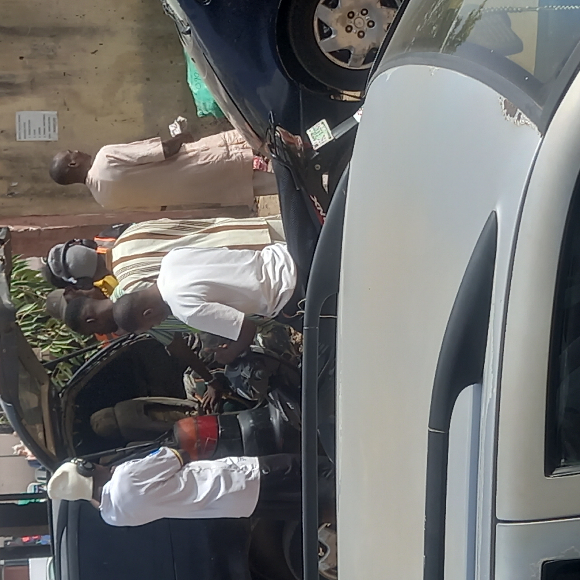 In Dadi Riba, Nigeria, a group of people are gathered around a car with its trunk open. The car appears to be parked on a street or roadside, with at least five individuals visible in the immediate vicinity. One man, wearing a white shirt and a white cap, appears to be near the trunk of the car. Two other men are looking into the trunk. Two additional men are standing away from the car. One man is in a pink robe. The car has a black exterior. The environment suggests an urban or populated area, given the presence of buildings and other vehicles.