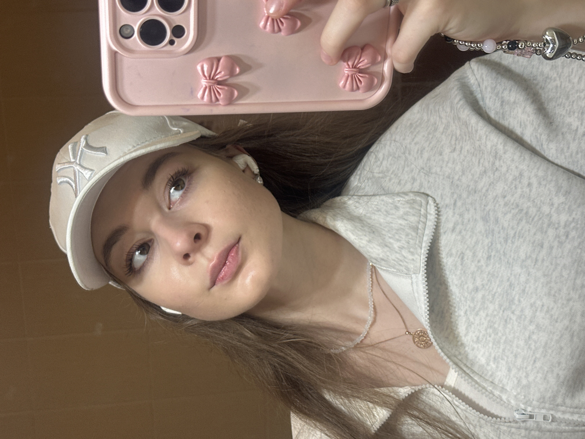 A young woman with long, brown hair, wearing a white baseball cap and a light gray zip-up hoodie, poses for a selfie. She is wearing a necklace with a circular pendant and another bracelet made of beads and a heart-shaped charm. Her phone, which has a pink case decorated with small bows and a camera cutout, is visible in the upper part of the image. The background is a plain brown surface. The lighting suggests it is daytime, possibly indoors. The woman's expression is neutral, and the overall mood is casual and relaxed. The setting appears to be a personal space, perhaps a home. There is no visible text in the image.