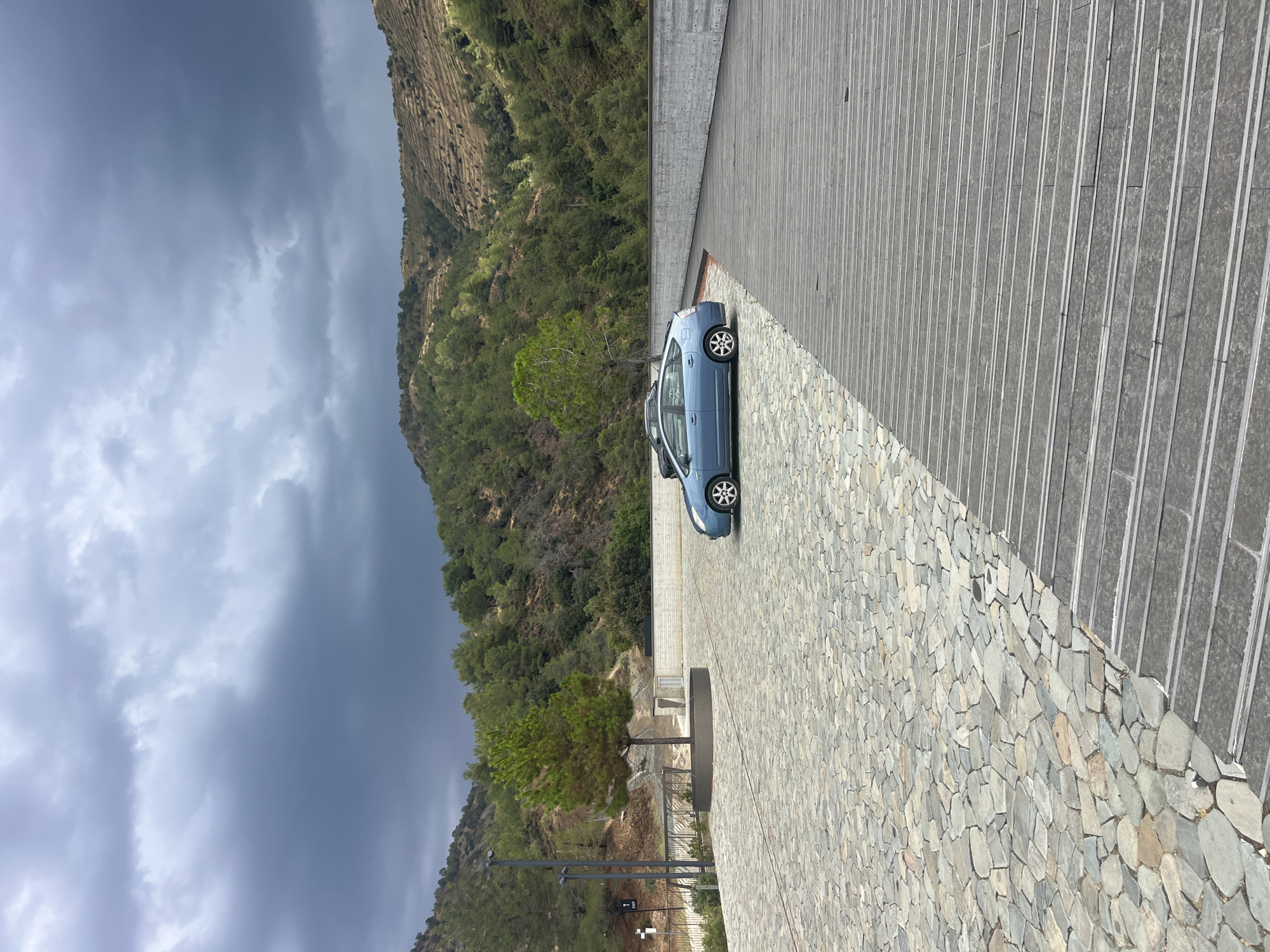The image depicts a parking area with a scenic mountainous backdrop in Kalopanagiotis, Cyprus. The foreground features a textured ground paved with irregular stone tiles, transitioning into a section of dark wooden planks arranged horizontally. Two cars are visible in the parking area: a blue hybrid sedan, possibly a Toyota Prius, parked in the center-left, and a dark-colored car partially visible behind it. Behind the parking area, a concrete wall runs horizontally across the frame. Beyond the wall, a wooded mountainside with terraced slopes is covered in lush green trees and vegetation. The sky above is heavily overcast with dark, dramatic storm clouds. A single mature pine tree stands prominently in a circular planter on the left side of the image, with a lamppost and an "EXIT" sign nearby. The overall atmosphere is somber due to the impending weather.