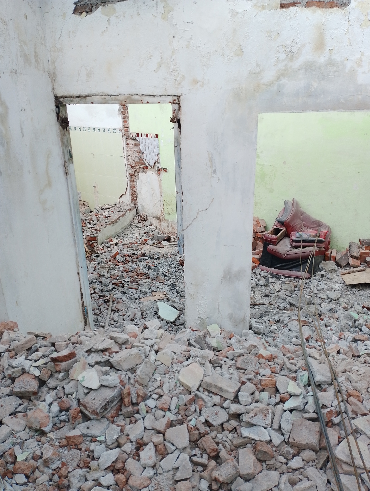 The image depicts a scene of demolition or construction debris within a building. The foreground is filled with a large pile of broken concrete, bricks, and plaster. The walls of the building are damaged, with peeling paint and exposed brickwork. In the background, through an open doorway, more debris is visible, as well as a light green painted wall. To the right, a red armchair is situated amongst the rubble, leaning against the light green wall. Two thin metal rods are also visible in the foreground, leaning against the debris pile. The overall impression is one of disarray and destruction. This scene is located in Payaman, Indonesia.