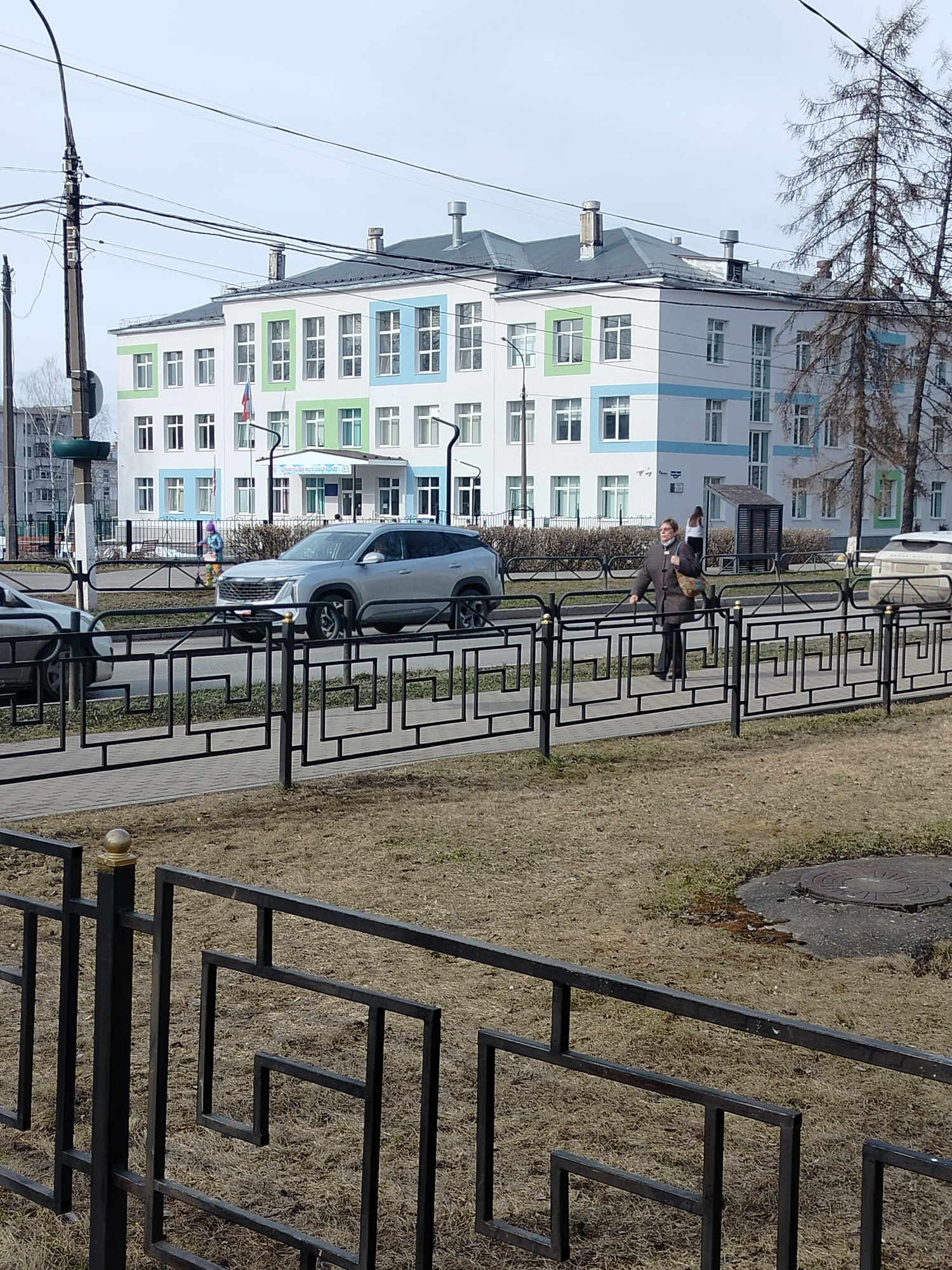 The image shows a street scene in the city of Chaikovsky, Russia. In the background is a three-story white building with blue and green trim around the windows. A silver SUV is parked on the street in front of the building, and a woman is walking along the sidewalk on the right side of the frame. A black metal fence runs along the sidewalk, and beyond the fence is a patch of dry grass. A gray utility pole is on the left, and other buildings and trees are visible in the background. The sky is overcast.