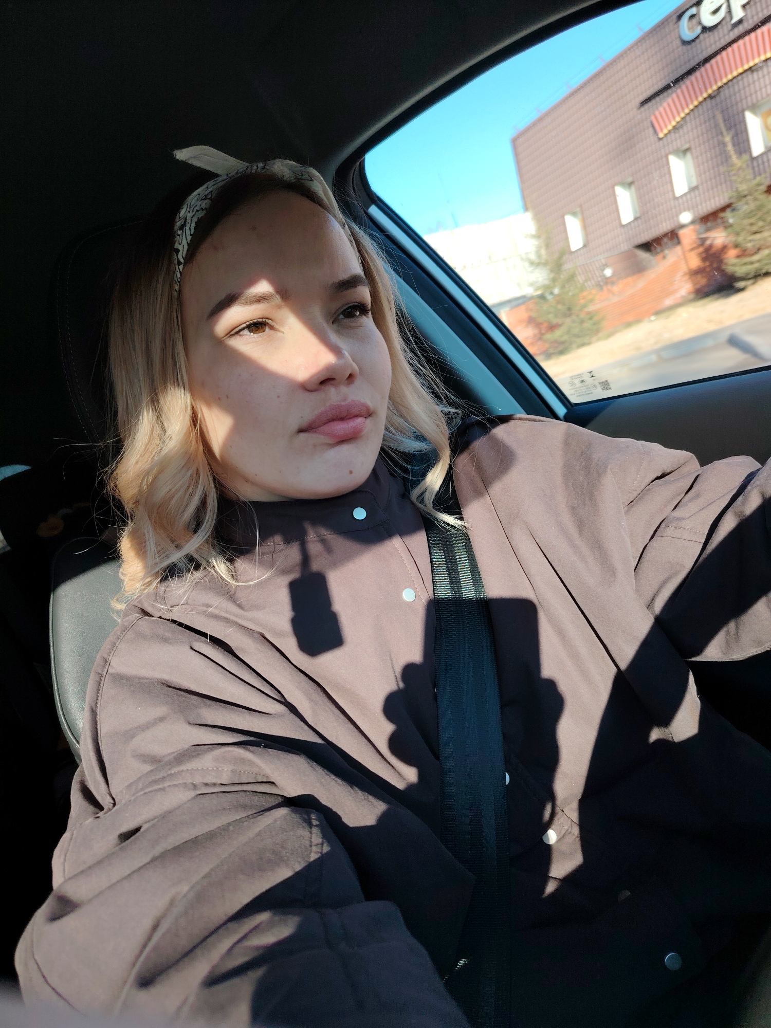 A young blonde woman with a headband is in the driver's seat of a car, wearing a brown jacket and a seatbelt. Sunlight casts shadows across her face and clothing. Outside the car window, a light brown building with a dark brown roof is visible, along with a clear blue sky. The building has a sign with Cyrillic letters, "ce," partially visible. Some greenery and red brickwork are seen near the building. The location is likely Chaikovsky, Russia, based on the building style and writing.