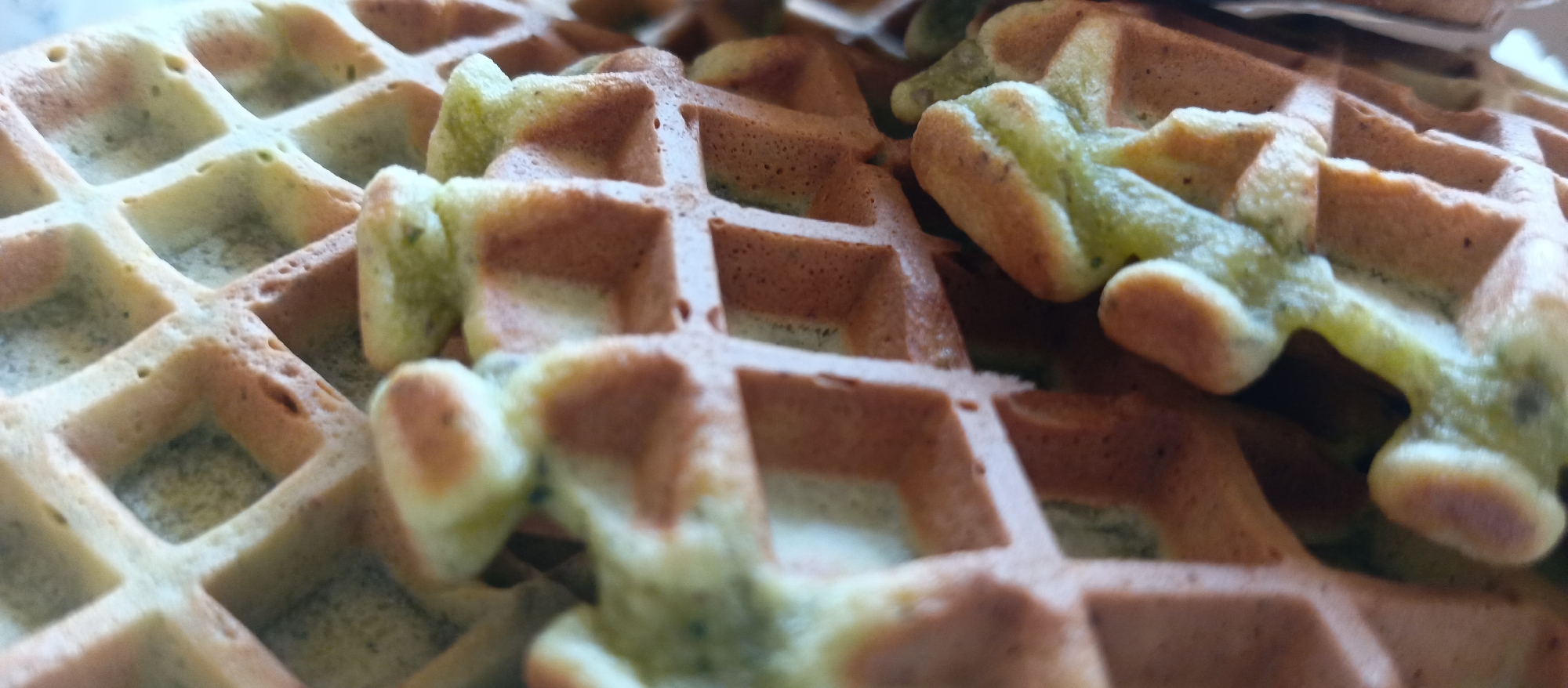 This close-up image displays a stack or pile of multiple waffles, filling the entire frame. The subjects are primarily these waffles, characterized by their distinct grid patterns.
A notable detail is the variation in color among the waffles. Some are a classic golden-brown, suggesting a traditional preparation, while others exhibit a light green hue, potentially indicating a specific flavor such as matcha or pandan. These green sections also show small dark specks, possibly remnants of the flavoring ingredients. The texture appears to be a mix of crispy edges and softer interiors, with the raised grids casting subtle shadows that add depth to the composition.
The photograph uses a shallow depth of field, with certain waffles in sharp focus while others in the foreground and background are softly blurred. This technique highlights the intricate details of the waffle closest to the viewer. There is no discernible activity or event taking place; the image is a static portrayal of the food item. No people are visible, and there are no visible texts or specific location cues beyond suggesting an indoor setting where food would be prepared or served. The time of day and weather cannot be determined from the image.