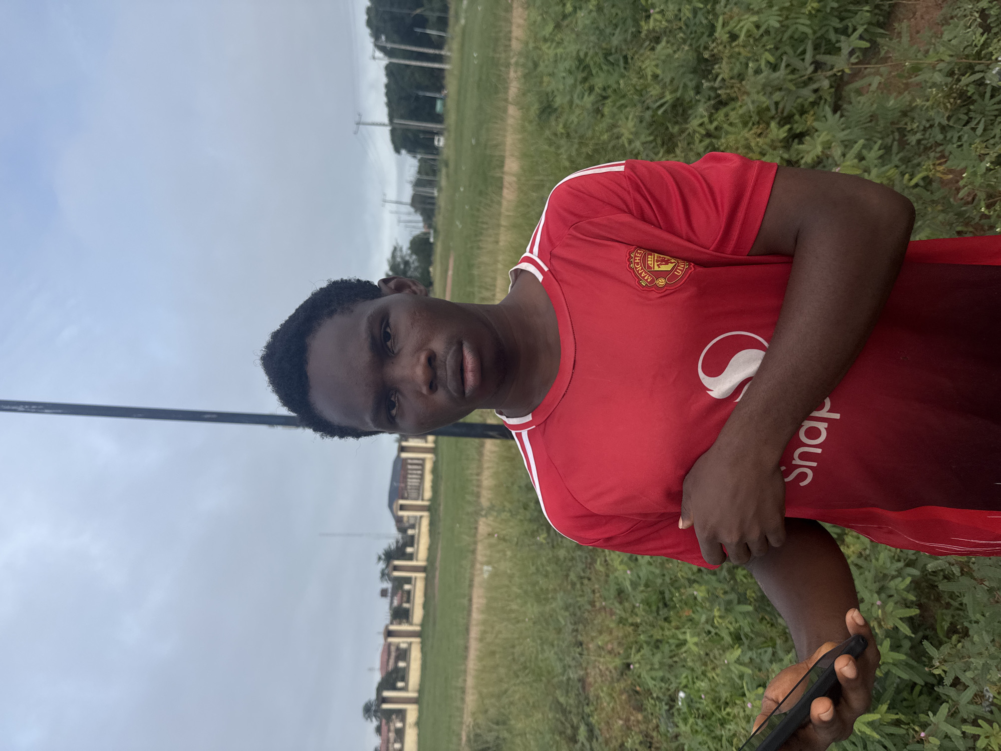 A young man in a red Manchester United soccer jersey stands in Ihiagwa, Nigeria. He is holding a black smartphone. Behind him are buildings, a grassy field, and utility poles under a cloudy sky.