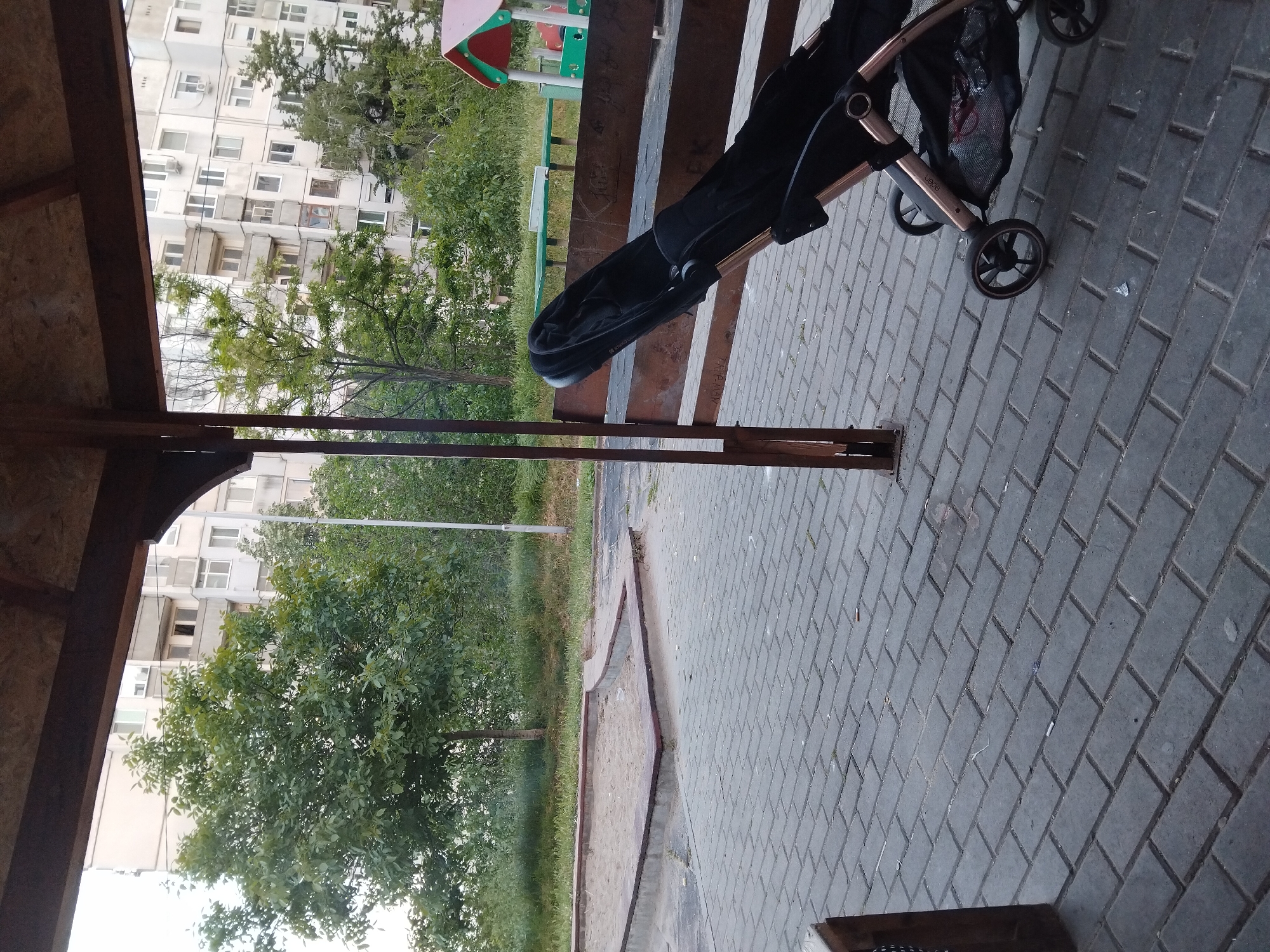 The image shows a scene at an outdoor location, likely in Stara Zagora, Bulgaria, featuring a baby stroller on a paved area next to a wooden structure, possibly a gazebo or shelter. The paved area consists of gray rectangular blocks, arranged in a herringbone pattern.
Beside the paved area is a sand pit bordered by concrete and grass. Beyond the sand pit, there are several trees and bushes, suggesting a park or green space. Further in the background, there is a multi-story residential building with numerous windows.
A baby stroller in black and rose gold tones is parked on the paved surface. The stroller has multiple wheels.
A playground area with colorful play equipment is visible in the background, slightly obscured by foliage.