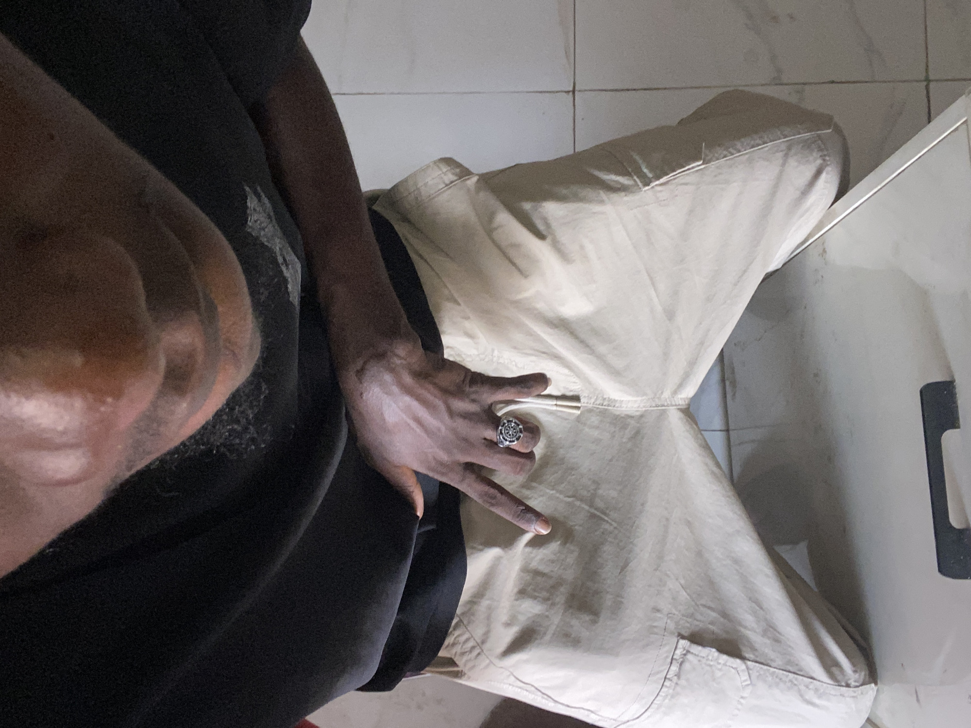 The image shows a person seated, wearing black clothing on the left and light tan pants on the right. The person's hand is visible, adorned with a large ring, resting on the pants near a drawstring. The background shows light-colored tiled flooring and part of what appears to be a white cabinet or wall with a handle. There is no definitive information in the image to confirm the location as Lagos, Nigeria. The description focuses only on the observable details within the photograph.