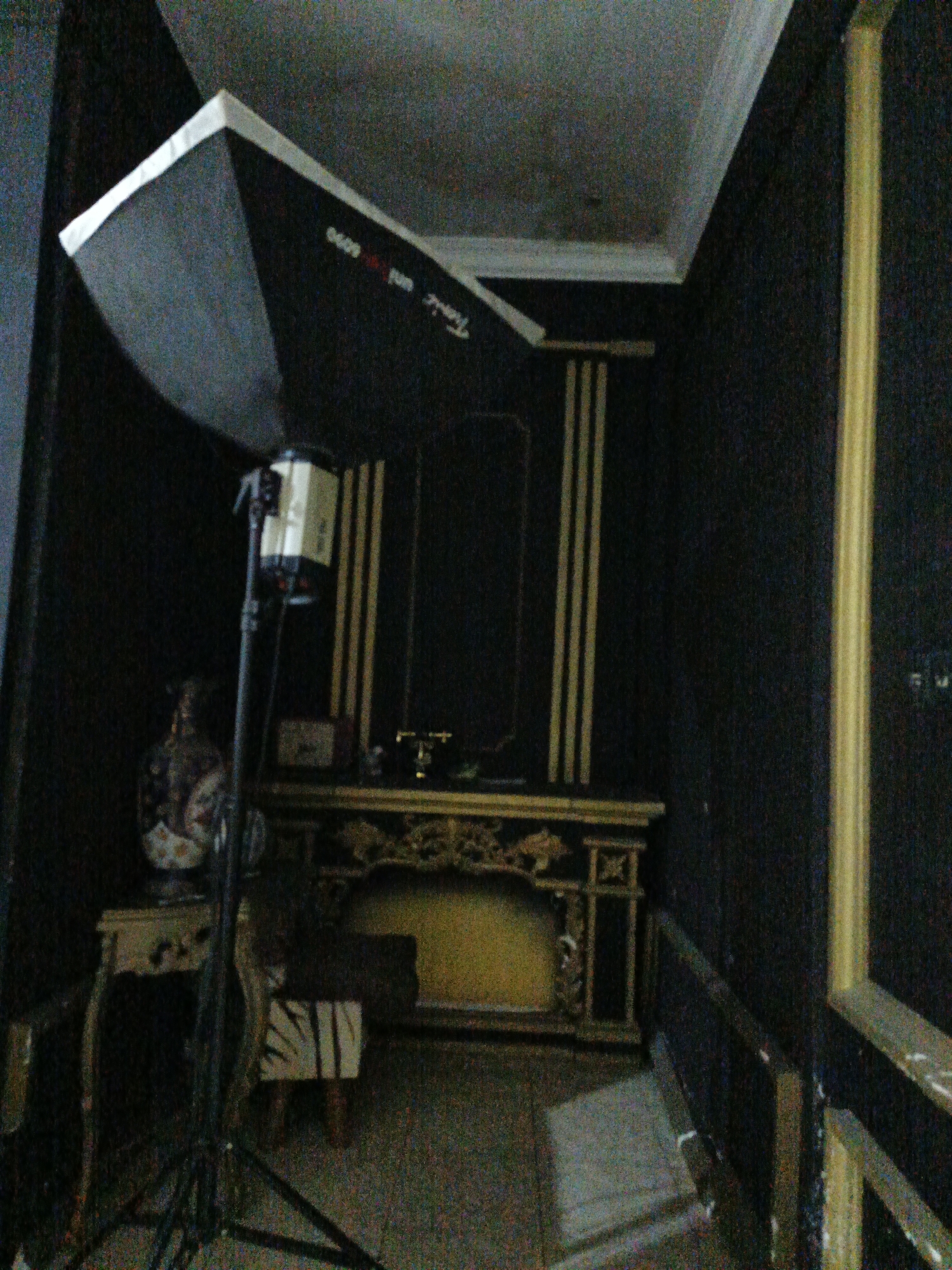 The image captures an indoor scene, possibly a studio or a room with dark, textured walls and decorative gold trim. In the foreground, a large softbox, likely part of photographic lighting equipment, is positioned on the left side of the frame. A light stand supporting a rectangular light fixture extends upwards from the softbox.
Dominating the center of the image is an ornate, dark-colored table or mantlepiece with elaborate gold detailing and carvings. It appears to have a yellow-lit recess in the center. To the left of this centerpiece, a small, ornate table with a carved leg supports a patterned vase or urn. A dark, indistinct object with a striped pattern rests on the floor in front of the main table.
The background reveals more of the dark walls, accented by vertical gold stripes. The ceiling is visible at the top of the image, with a lighter textured finish. The overall impression is of a dimly lit, intentionally styled environment, possibly set up for photography or a decorative display. The location is identified as Karangpakel, Indonesia.