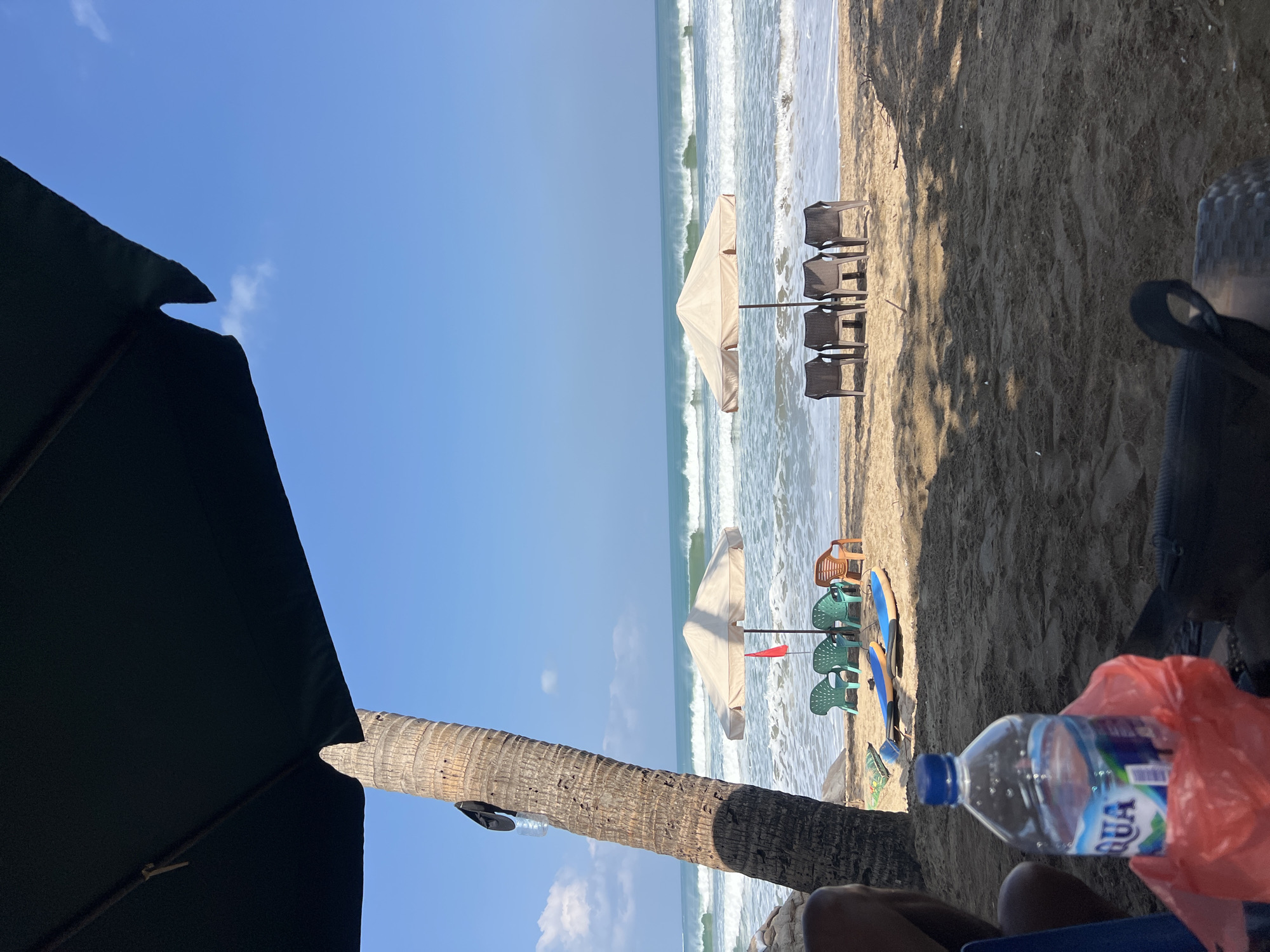 This is a medium shot, eye-level, outdoor photograph of a beach in Kuta, Indonesia. The image is dominated by a large black umbrella in the foreground, casting a shadow over a portion of the sandy beach. To the left of the frame, a palm tree trunk rises vertically, with a plastic water bottle and a black item hanging from it.
In the middle ground, two beige beach umbrellas stand on the sand, each with chairs arranged in front of them. The umbrella on the left has four green plastic chairs and one orange plastic chair. A red flag is planted near this umbrella. Two surfboards, one blue and one orange, are laid out on the sand in front of these chairs. The umbrella on the right has four brown plastic chairs.
The background features the ocean with gentle waves breaking and a clear blue sky with scattered white clouds. The sand appears damp near the water's edge. The overall scene depicts a tranquil beach setting with amenities for relaxation and recreation.