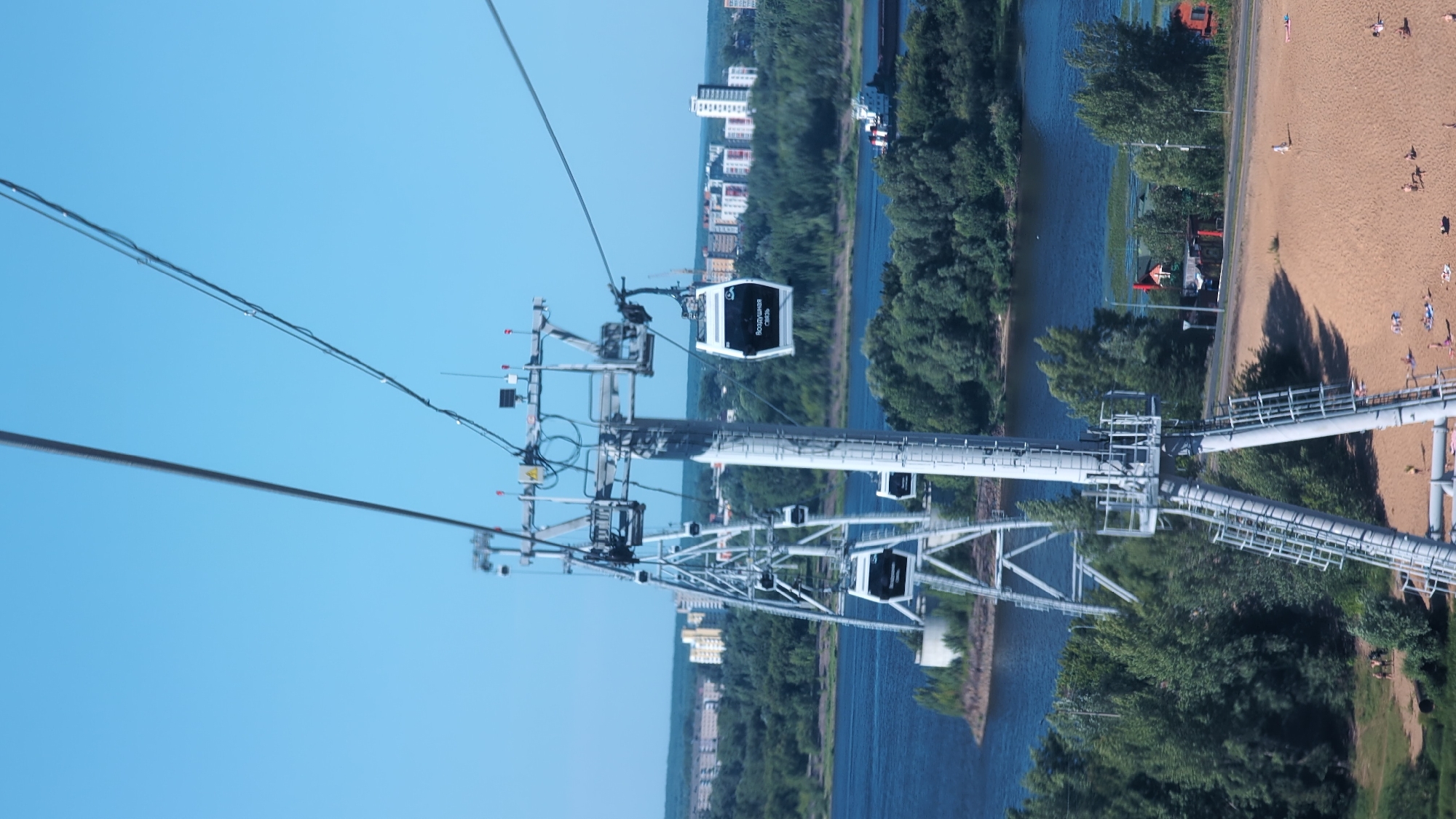 The image captures a view from a cable car, showing a wide blue sky and a cable car suspended over a river. Several cable car cabins are visible, some attached to the cable, others on the support structures. The support structures themselves are large and metallic.
Beyond the river, a lush green forest lines the bank, with a sandy beach populated by several people scattered across it, some sunbathing or walking. Further inland, buildings, including apartment complexes and cranes, suggest an urban or suburban landscape. The river is a dark blue, reflecting the sky.
The perspective is from the cable car, looking downwards and across the scene. The time appears to be daytime, with bright sunlight casting shadows. The weather seems clear and pleasant.
One cable car cabin has Cyrillic text on its side, reading "Роудюная Окно", which translates to "Roadway Window". The overall impression is one of a scenic view from a recreational cable car ride, likely in Nizhny Novgorod, Russia, given the location context.
