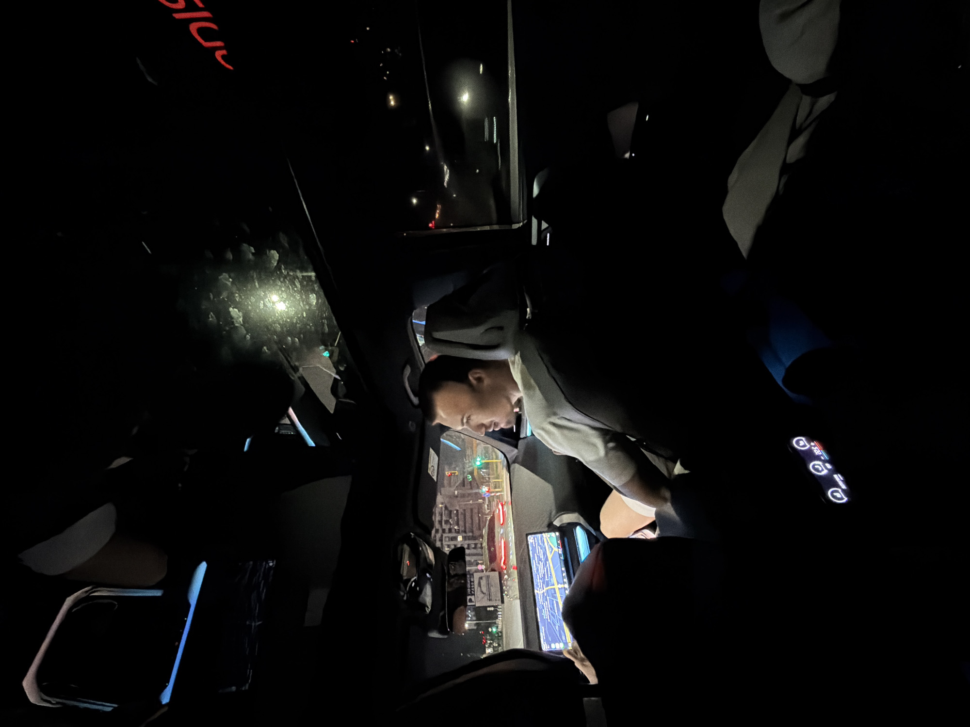 The image displays the interior of a vehicle at night, situated at an urban intersection in Sofia, Bulgaria. The car's expansive digital navigation screen clearly shows a map of Sofia with Cyrillic street names, specifically identifying "бул. България" (Bulgaria Blvd) and "бул. Петко Каравелов" (Petko Karavelov St.), indicating the vehicle's position.
Two individuals are present in the front. The driver is partially visible, with their legs in light-colored shorts and one hand positioned near the steering area. The front passenger is a male, estimated to be in his 20s or 30s, wearing a light-colored short-sleeved t-shirt and light shorts. He is secured by a visible seatbelt and is looking towards the left, potentially at the driver, the navigation screen, or the passing streetscape.
The car's interior is modern, featuring a wide, integrated digital display that functions as both the instrument cluster and infotainment system. Below this primary screen, a smaller panel with illuminated touch controls is visible. Blue ambient lighting accentuates various elements of the car's cabin. A panoramic sunroof is visible above, showing reflections and what appears to be condensation or water droplets.
Through the windshield, a nighttime city environment is observed, characterized by illuminated buildings, active traffic lights (showing both red and green signals), and other vehicles, including a red car at the intersection. A partial red text reflection, possibly "2019" or "DIG", is visible on the upper right portion of the windshield/sunroof. The passenger-side window reflects another vehicle. A grey garment is folded and resting on the rear seat or console area on the right side of the vehicle.
