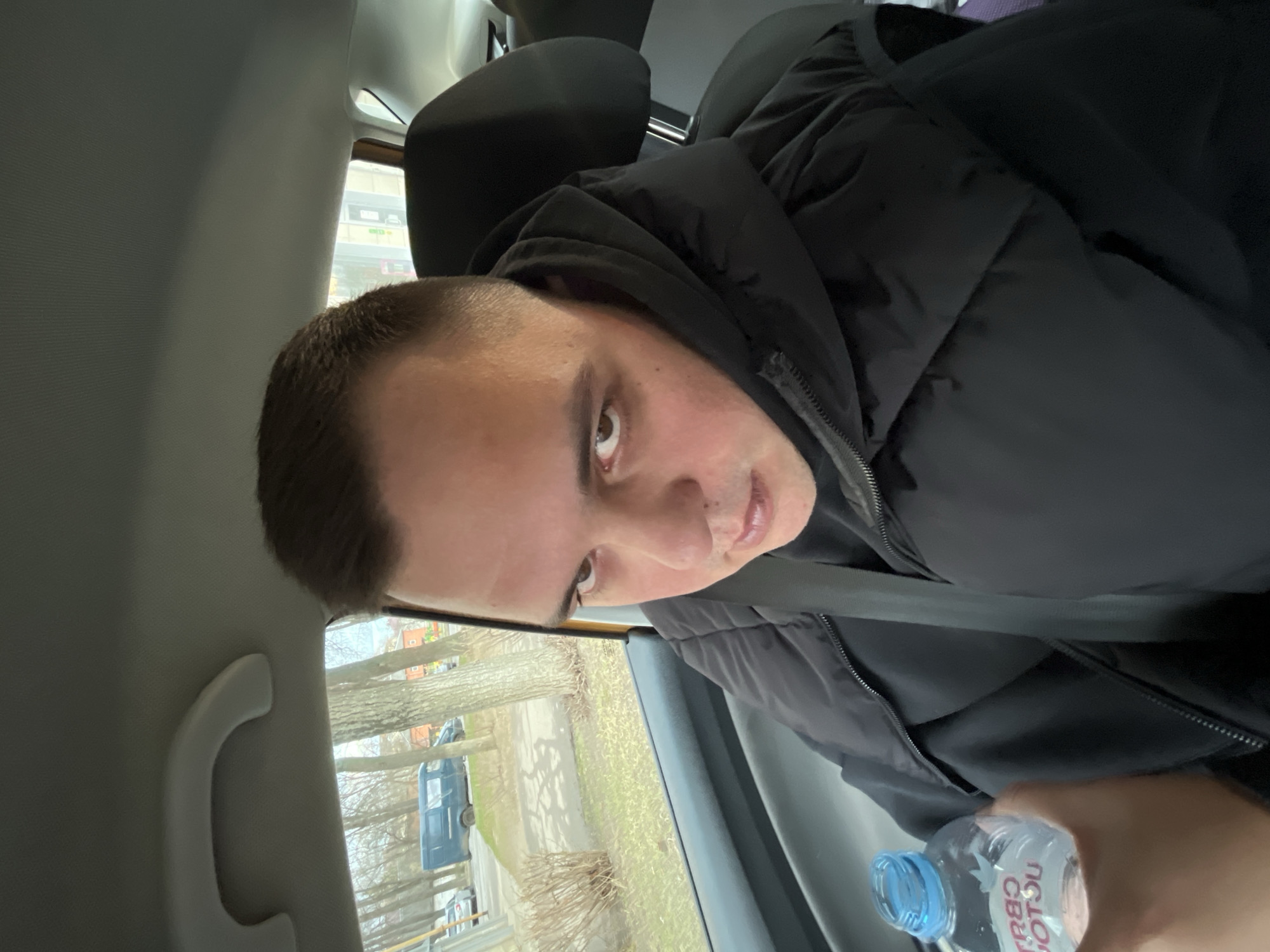A man is seated in the passenger seat of a car, wearing a black hooded puffer jacket and a seatbelt. He is holding a bottle of water in his right hand. Outside the car window is a view of a street in Klin, Russia, with bare trees and parked vehicles. The window shows some buildings. The sky appears overcast. The man is looking at the camera with a serious expression. The car's interior is visible, including the headrest, the door handle, and the grey interior paneling.