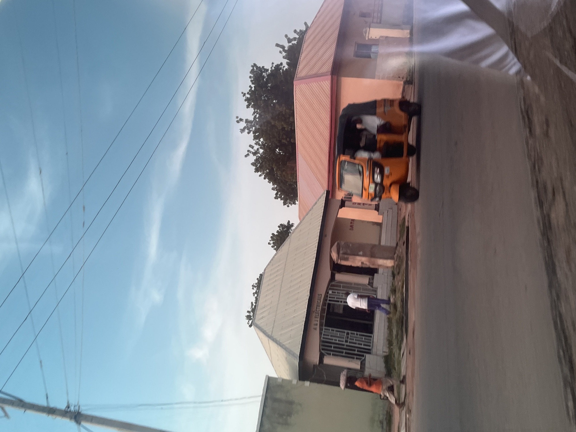 A yellow auto-rickshaw is parked on the side of a road in front of two buildings, one with a pink roof and the other with a grey roof. The pink-roofed building has a window and a door, and the grey-roofed building has an entrance with bars. Two people are visible; one is standing by the grey-roofed building, and another is sitting in the auto-rickshaw. The scene appears to be set in a town or city, with power lines visible in the sky. The sky is a light blue with some white clouds, suggesting it is daytime. The overall impression is a snapshot of everyday life in a Nigerian community. The auto-rickshaw, commonly known as a 'Keke' in Nigeria, is a popular mode of transportation in Jalingo. The buildings seem to be shops or businesses, with one possibly being an electrical shop, as indicated by the text "A M J ELECTRICALS" on the grey building. The time of day is likely late afternoon or early evening, given the lighting.
