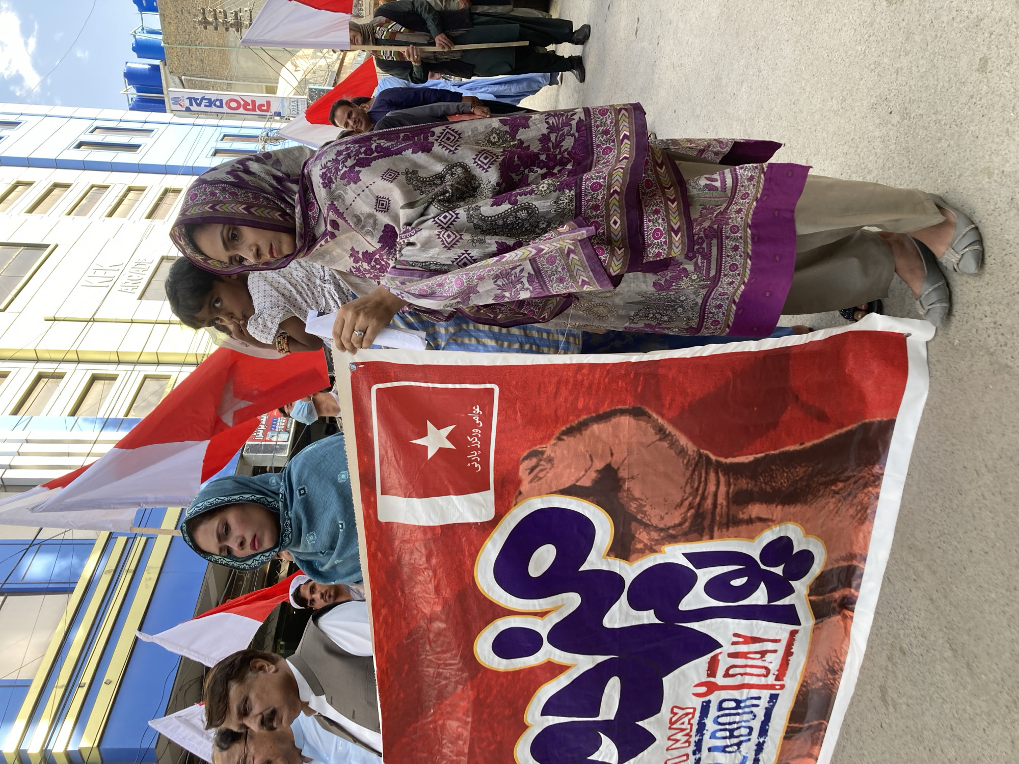 A street scene in Quetta City Tehsil, Pakistan, features a group of people holding a banner. The banner, primarily red and white, has a white star and writing in an unknown language. The text "LABOR DAY" is also visible. A woman wearing a patterned purple and beige hijab and outfit stands prominently near the banner. Other individuals are behind the banner, some holding red and white flags. Buildings are in the background with the sign "PRO DEAL" visible on one. The sky is light blue. The banner obstructs the view of a majority of the bodies of the protesters; several only reveal the top of their head.