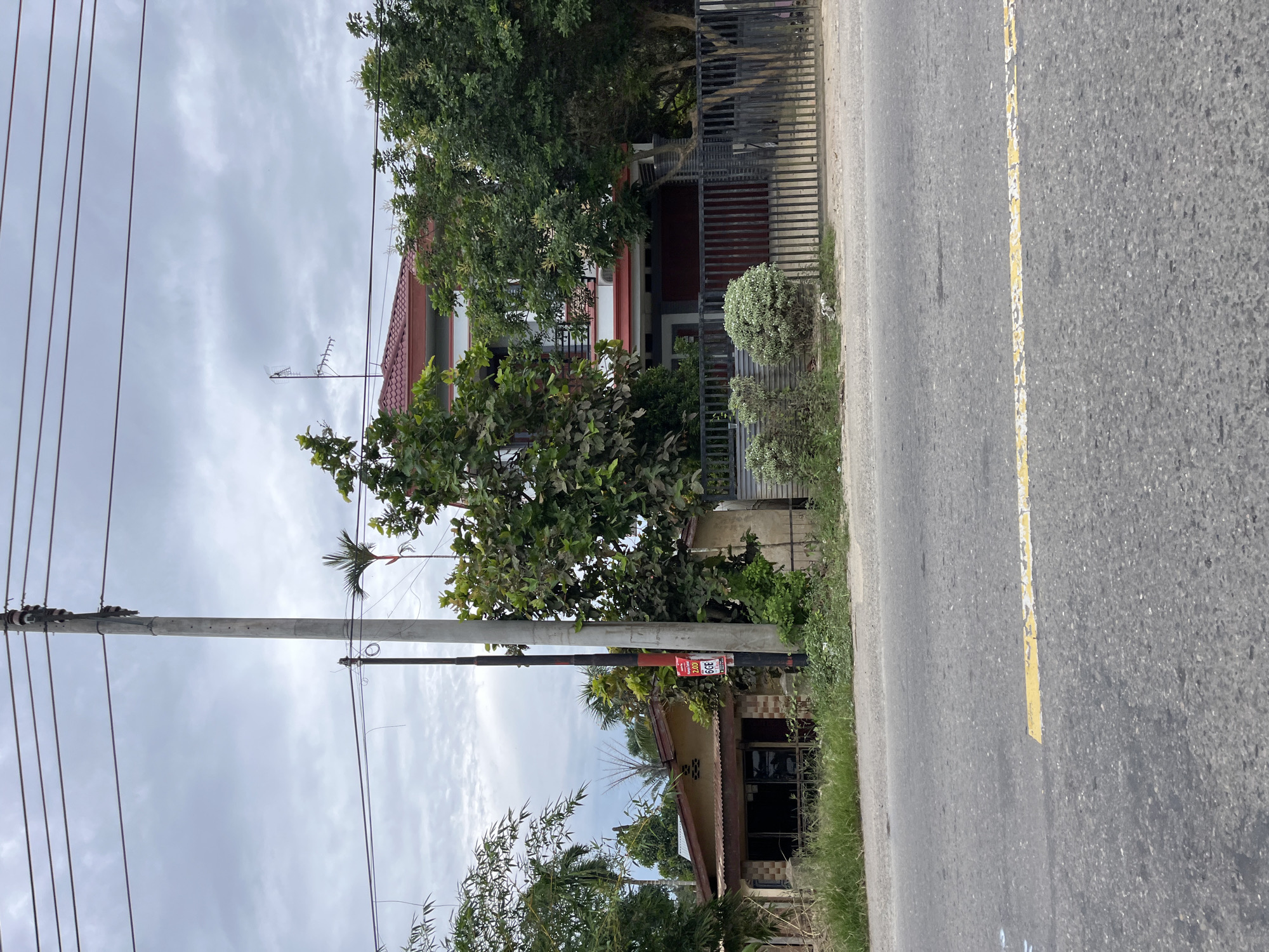 The image depicts a street scene in Simpangempat, Indonesia, under an overcast sky. A paved road with a faded yellow line runs through the foreground. To the right of the road, a tall, gray concrete utility pole stands, adorned with red and black banding, and supporting multiple electrical wires. Behind the pole, a two-story house with a red roof and white trim is visible, partially obscured by lush green trees. The house has an attached garage with a dark red door and a black metal fence in front. To the left of the utility pole, a smaller, single-story building with a corrugated metal roof and a covered porch is situated. This building appears to have large windows with dark frames and is surrounded by overgrown grass and vegetation. A red and white sign with text is attached to the utility pole. The overall environment suggests a semi-urban or suburban setting. There are no people visible in the image.