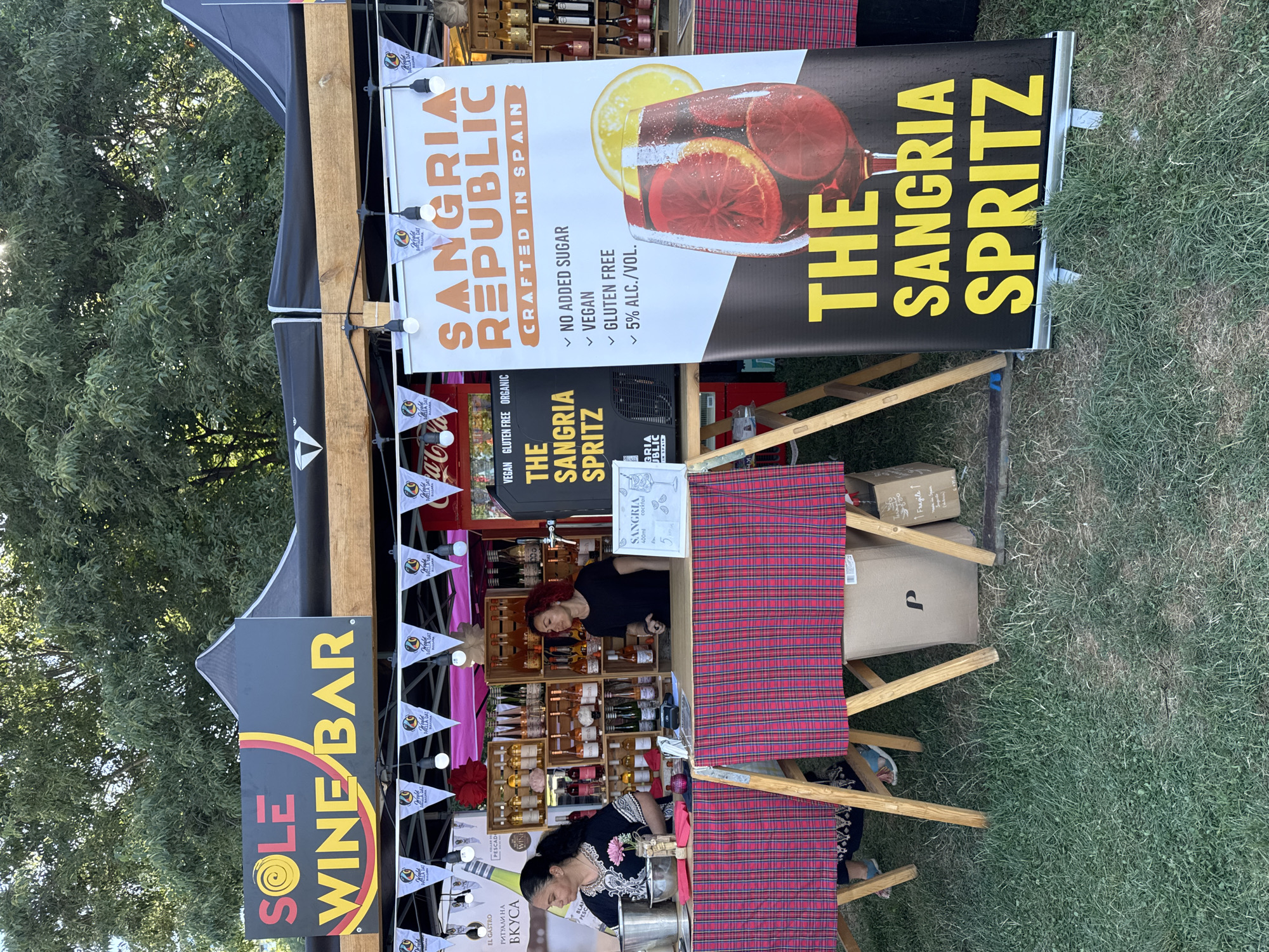 The image depicts an outdoor stall selling wine and sangria spritz in Burgas, Bulgaria. The stall is branded as "SOLE WINE BAR" and features a large banner for "SANGRIA REPUBLIC CRAFTED IN SPAIN." Two women are working at the stall. One woman, on the left, is wearing a black and white patterned dress and is preparing drinks. The other woman, on the right, is wearing a black t-shirt and is standing behind the counter. Various bottles of wine are displayed on shelves behind the counter. The stall is decorated with small triangular flags and string lights. In the foreground, a red and black plaid tablecloth covers the tables. Boxes are visible underneath the tables, and the ground is covered in grass. The background shows trees and a tent structure.