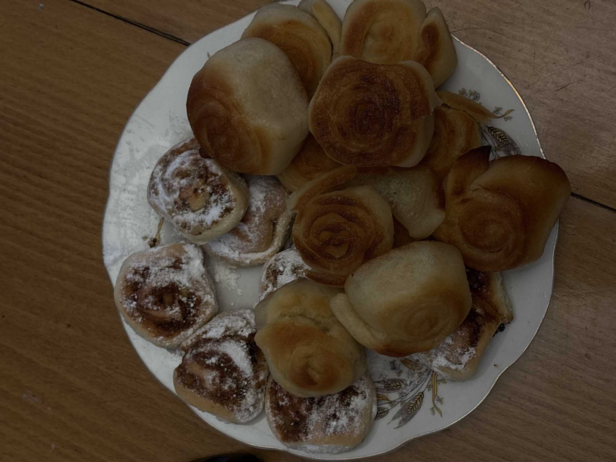 A plate of pastries sits on a wooden surface. Some pastries are round and sprinkled with powdered sugar, revealing a brown swirl pattern. Other pastries are smooth, golden-brown rolls. The plate itself is white with a decorative gold-colored pattern around the rim. The image is taken from an overhead perspective. There is no indication of a location in Borino, Bulgaria.