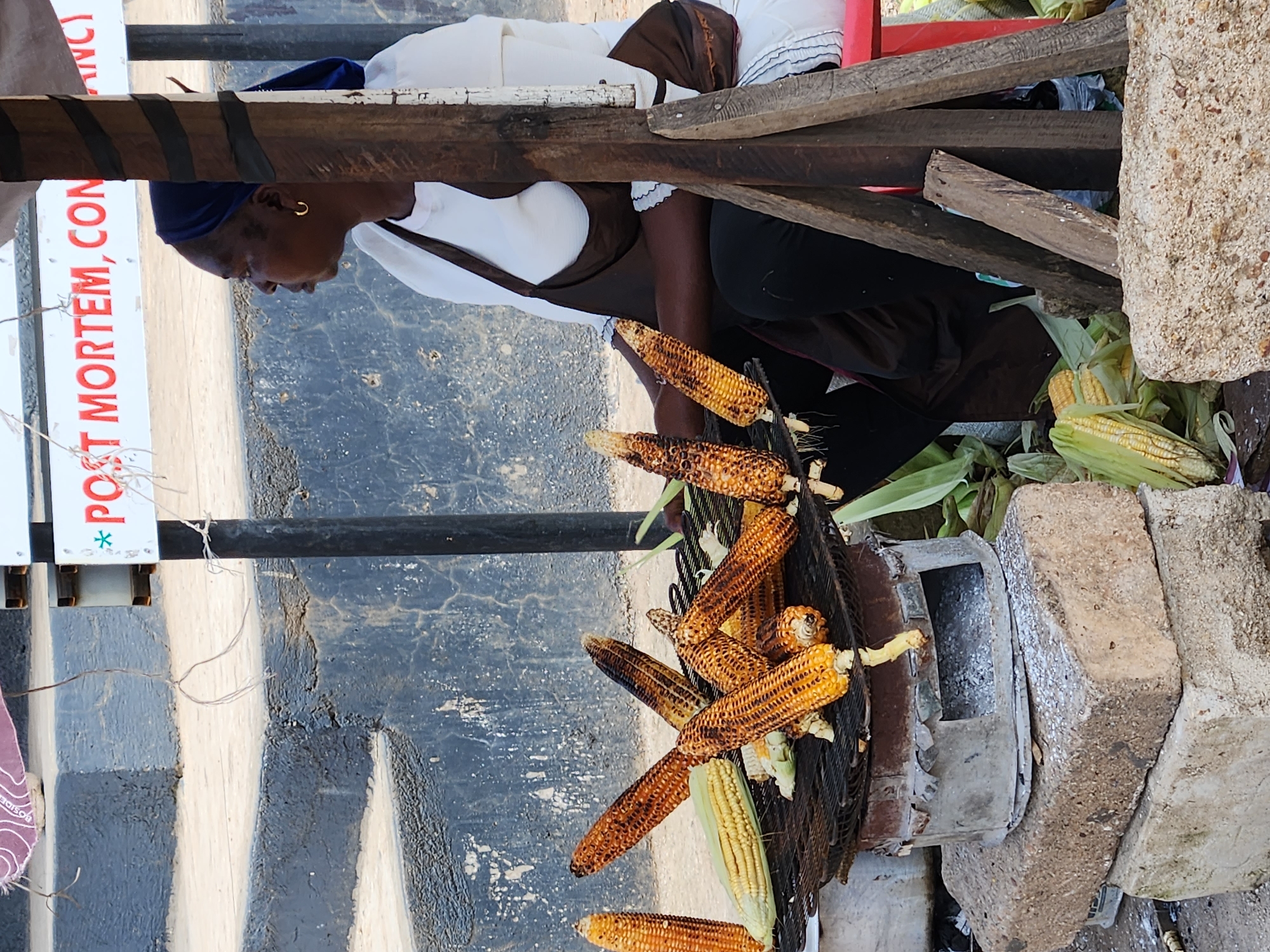 A woman wearing a dark blue headscarf and a white top with a brown apron is sitting behind a stand selling grilled corn. The stand is constructed from wooden planks and has a metal grill over a charcoal stove that sits on stacked concrete blocks. Several ears of corn, some grilled and some raw, are visible on the grill and around the stand. A sign with red lettering is visible in the background, reading "POST MORTEM, CON". The scene appears to be outdoors, possibly on a street in Jos, Nigeria, given the cultural context of street food vendors. The woman is looking to her right, away from the camera, with a neutral expression. The environment suggests a typical street market setting with a worn concrete wall in the background and some debris on the ground.