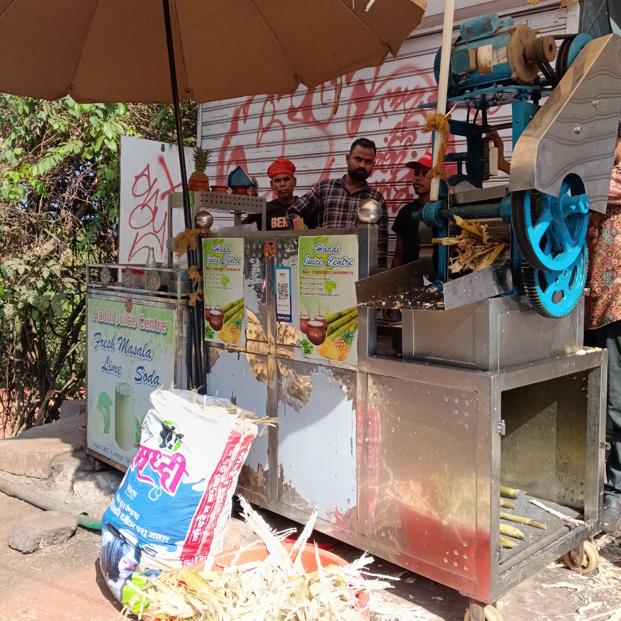 The image shows a "Haddi Juice Centre" stall in Mapusa, India. Three men stand behind the stainless steel stall. One wears a red turban and a black t-shirt with "BEK" printed on it; another wears a checkered shirt; and the third wears a red baseball cap and a black t-shirt. The stall has signage advertising "Fresh Masala Lime Soda" and "Sugarcane Pineapple Mix Juice." A blue sugarcane juice extractor is attached to the right side of the stall, with sugarcane stalks visible in the machine. A bag labeled "Samruddhi" with an image of a cow on it leans against the stall, and a container filled with sugarcane remnants sits in front of it. The stall stands under a large brown umbrella. The background includes a closed storefront covered in graffiti and some greenery.