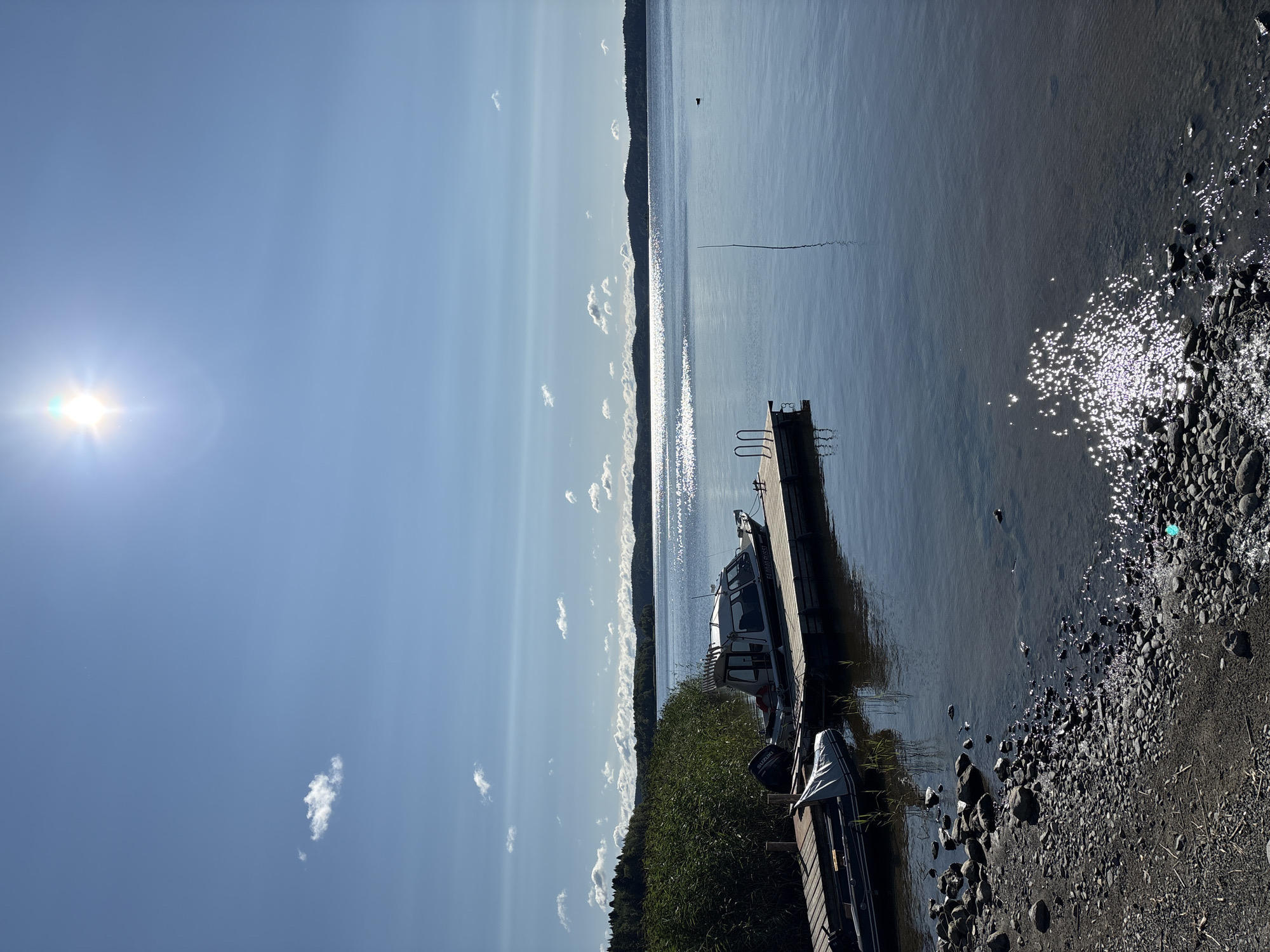 The image depicts a serene lakeside scene at Rautalakhti, Russia, under a bright, sunny sky with scattered clouds. A wooden dock extends into the calm, clear water, where two boats are moored. One is a larger cabin boat, white with a dark stripe, and the other is a smaller inflatable boat. The shoreline is covered with pebbles and dark earth, with tall reeds growing along the edge. In the distance, the lake stretches to meet a tree-lined horizon of rolling hills, bathed in sunlight that creates a shimmering path across the water's surface. A single dark object, possibly a bird, is visible on the water in the middle distance. A thin pole stands vertically in the water to the right of the dock. The sun is high in the sky, casting a bright glare.