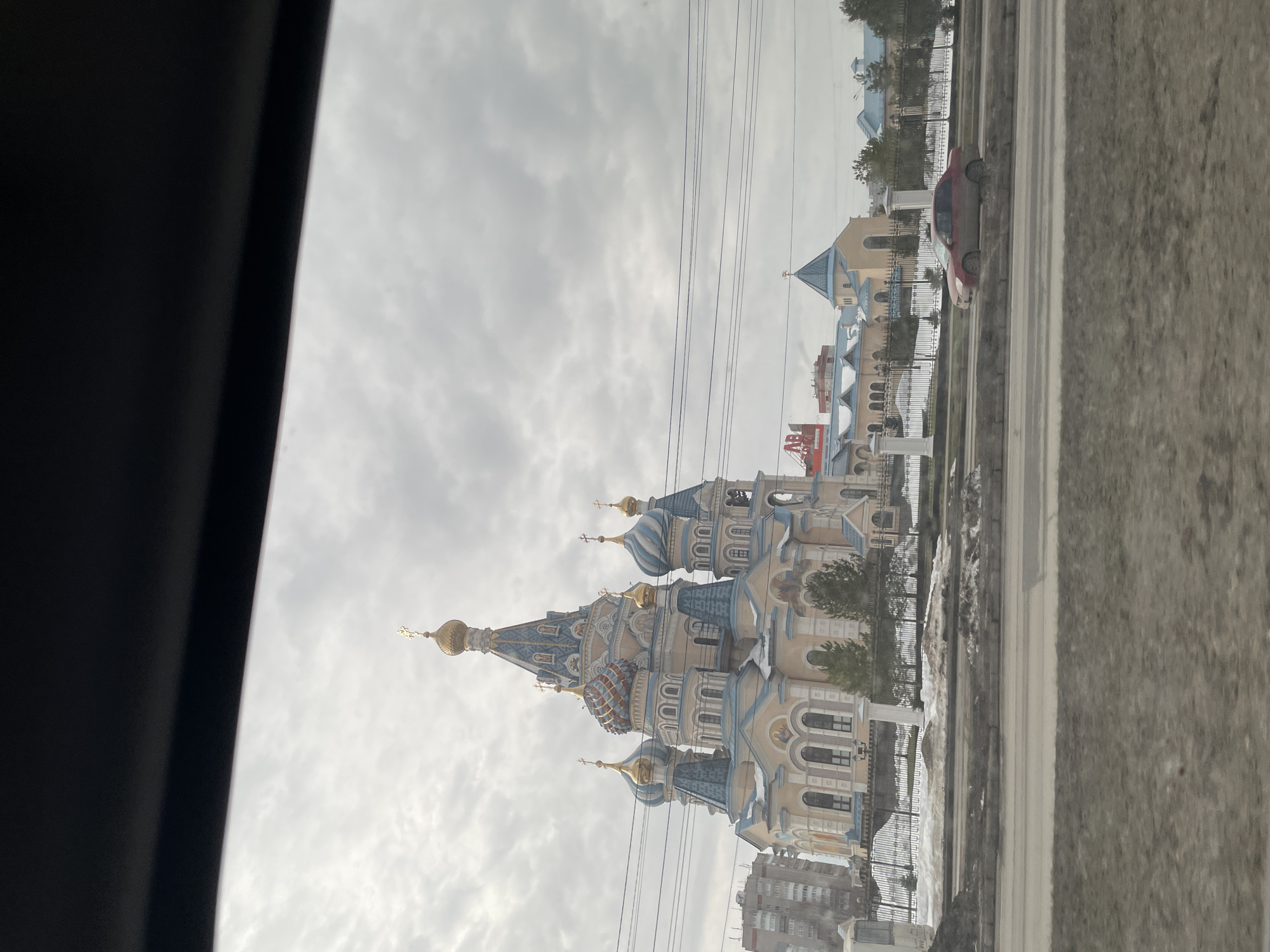 The image shows the Alexander Nevsky Cathedral in Izhevsk, Russia. The cathedral features golden domes and light blue and white exterior details. The sky is overcast. In the foreground are the interior of a vehicle and an outdoor view of a cityscape on a cloudy day. There is also a view of a road with a car parked on the side.